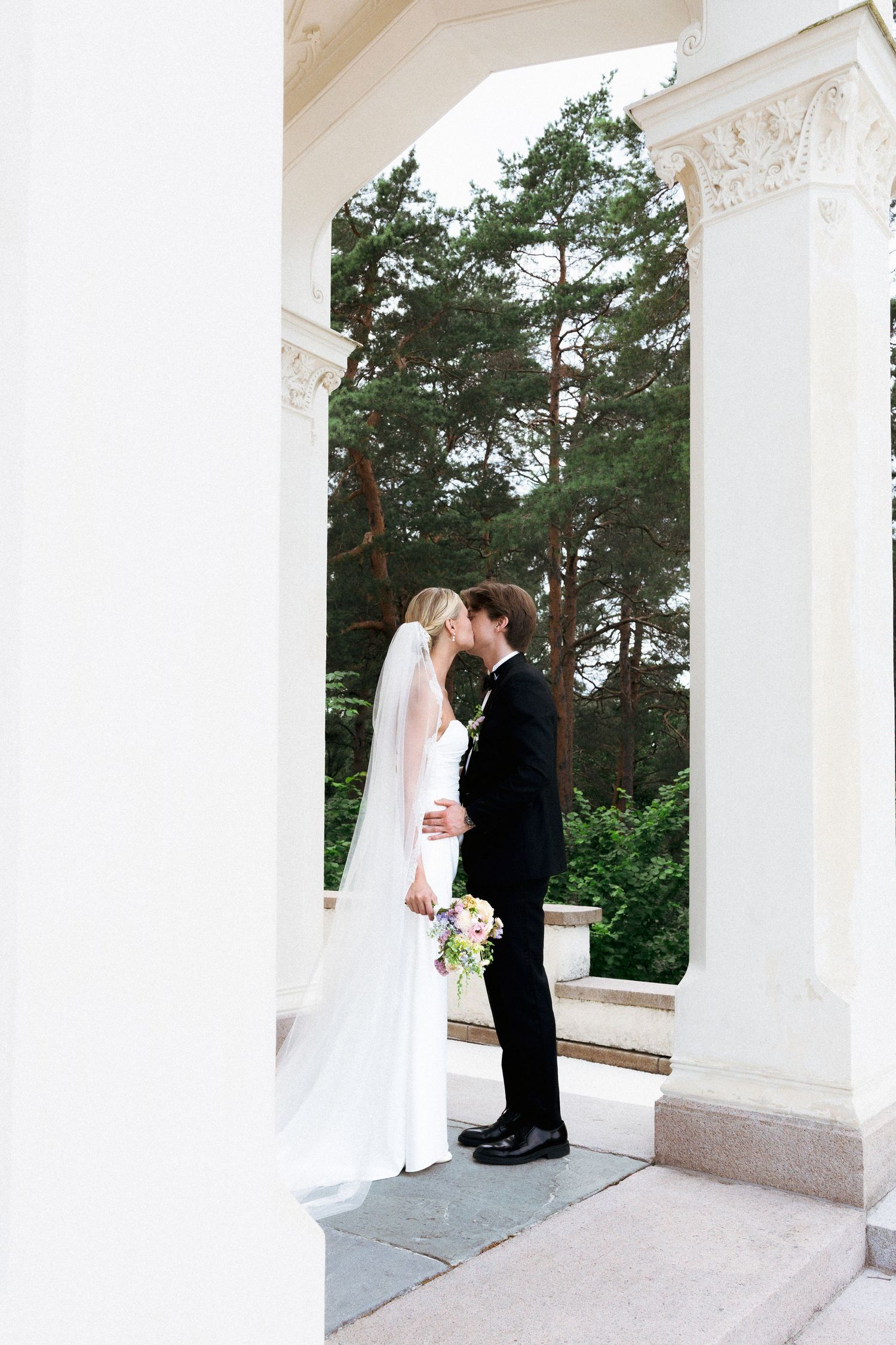 Bride and groom sharing a kiss between white columns with lush green trees in background.