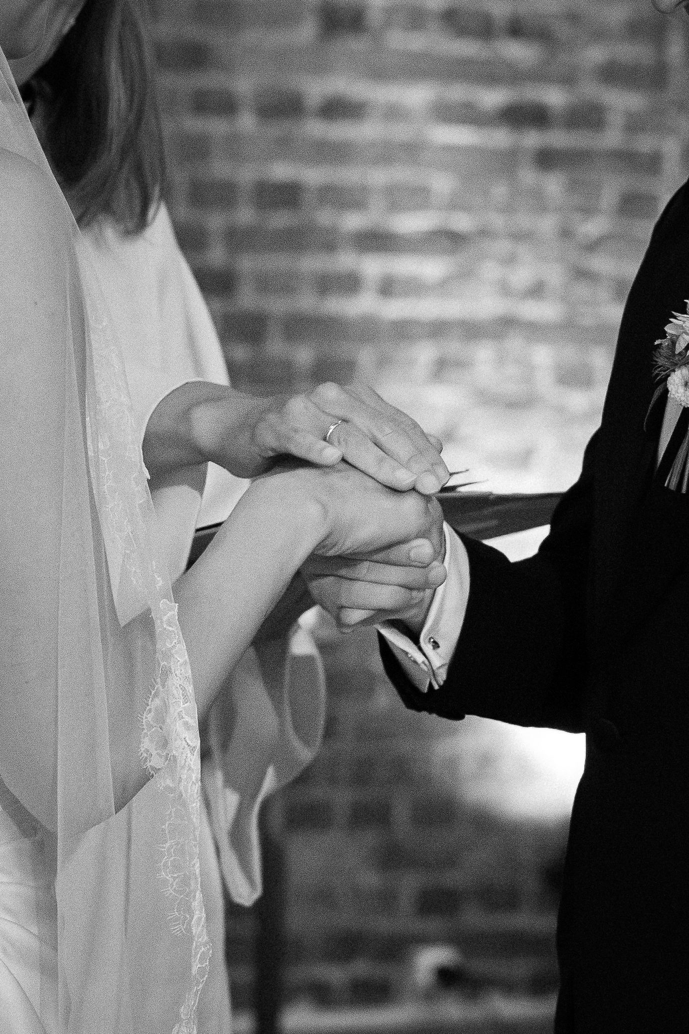 Black and white photo of bride and groom exchanging wedding rings during ceremony near brick wall.
