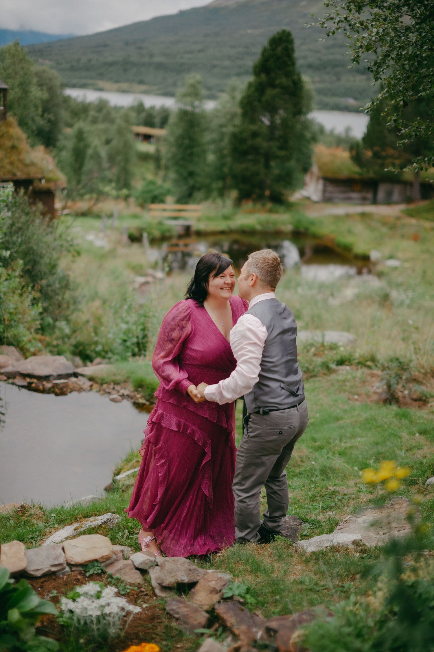 Couple in love dancing outdoors near a pond, woman in burgundy dress, man in gray vest, lush green mountain backdrop.