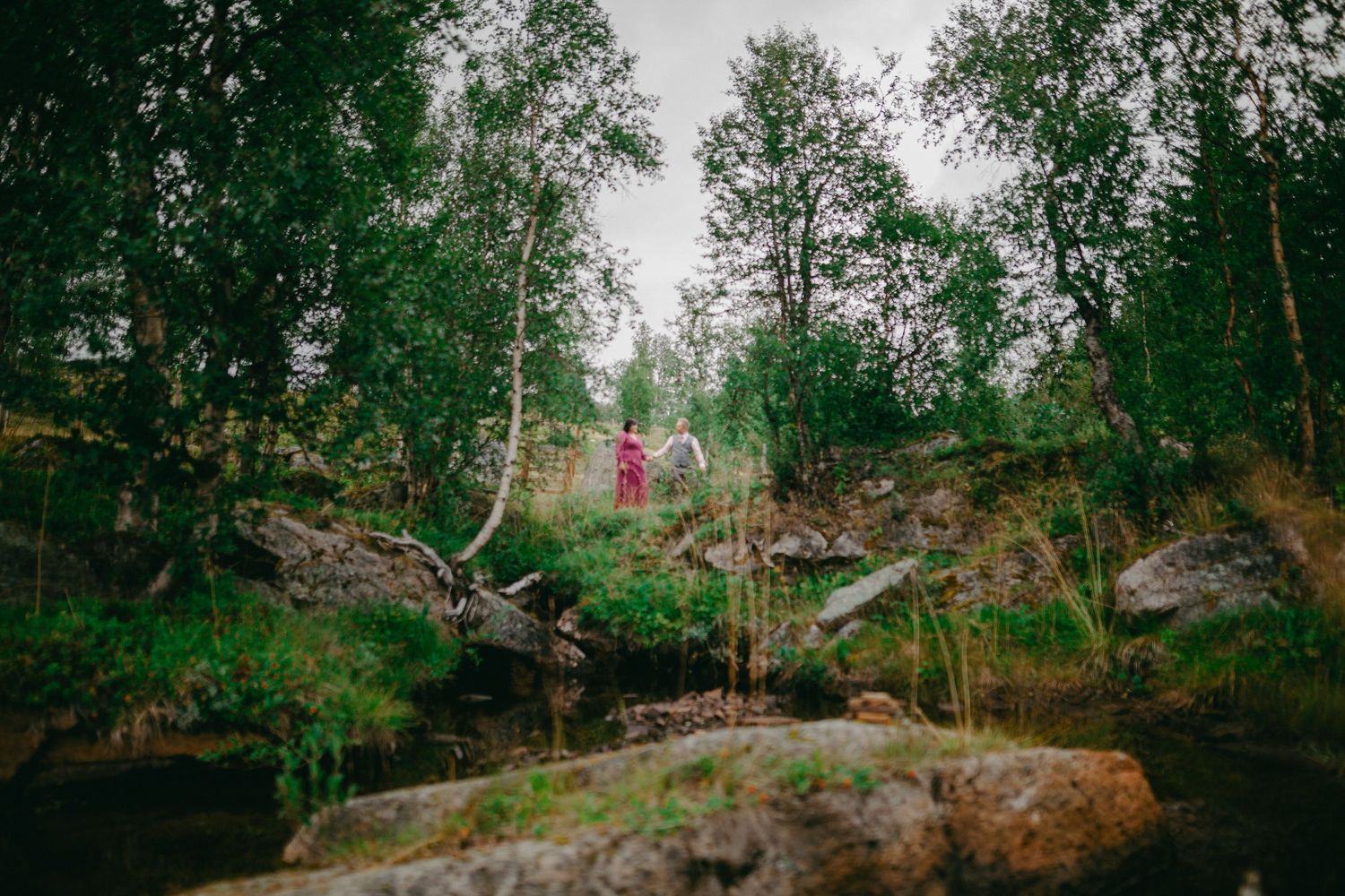 Person in red standing among lush green trees and mossy rocks beside a small stream in a forest.