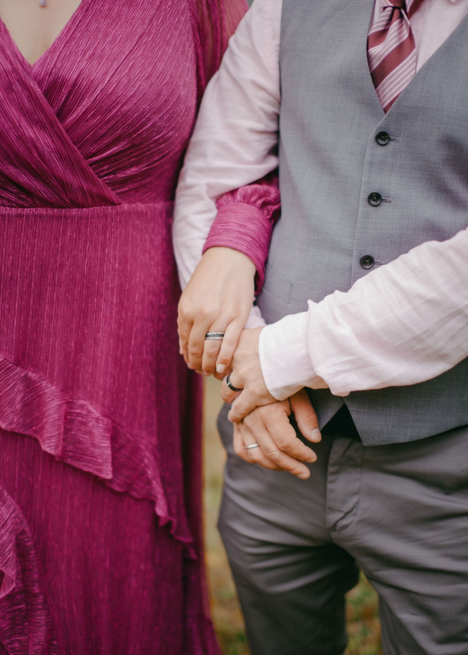 Couple holding hands, woman in magenta dress with diamond ring, man in gray vest and striped tie.