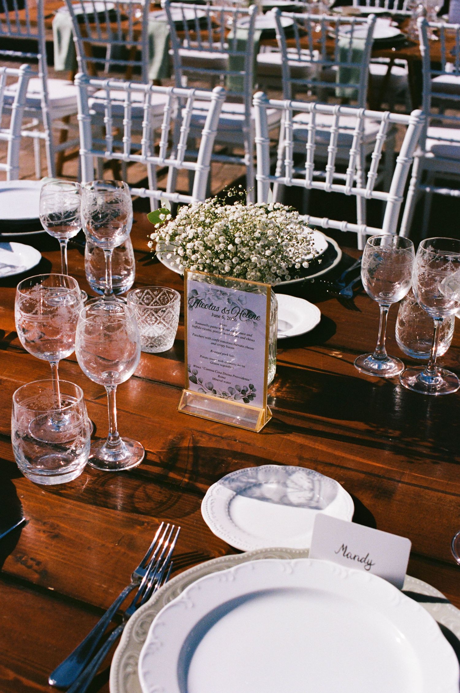 Elegant wedding reception table set with crystal glasses, white floral centerpiece, place cards, and white china on a wooden table.