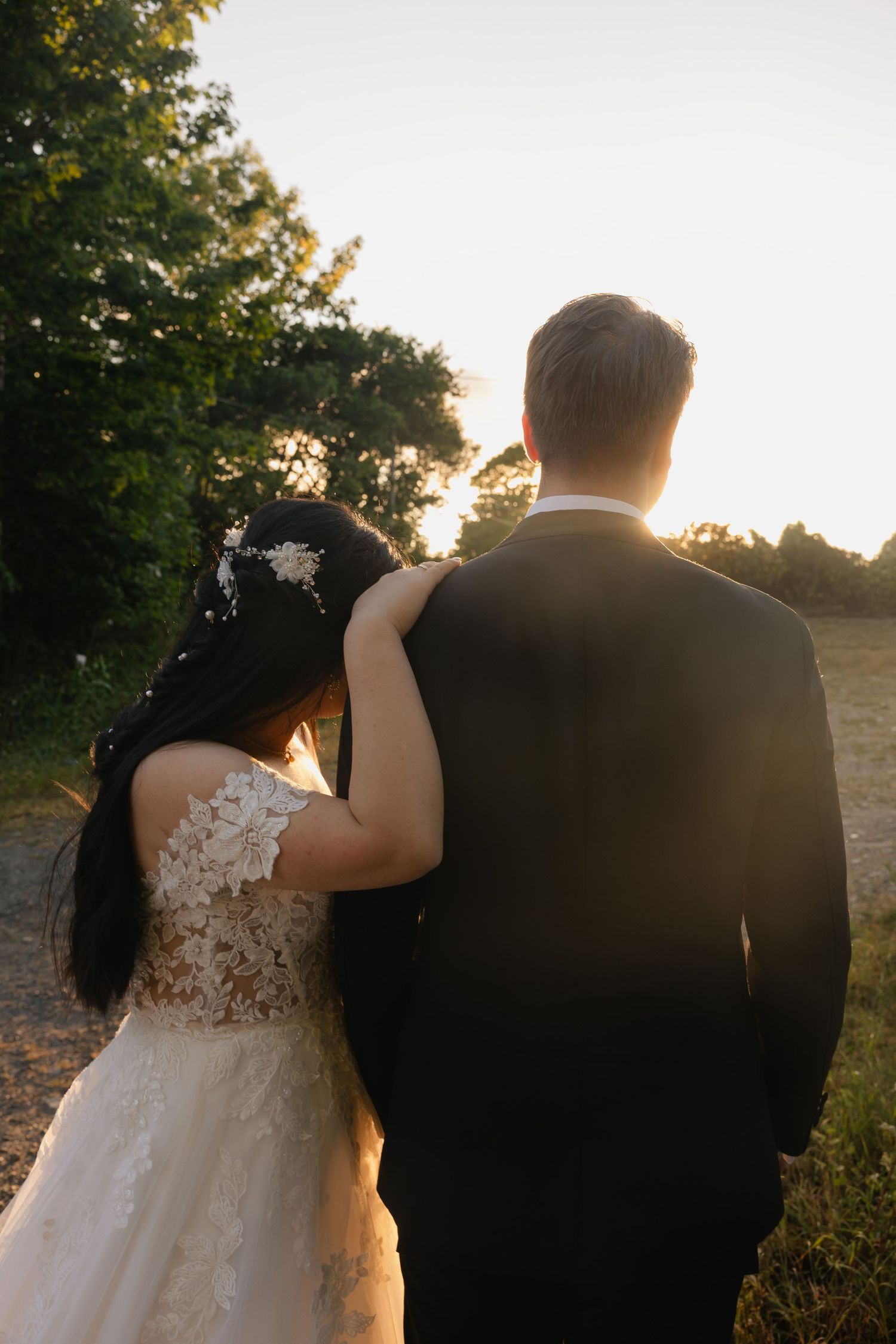 Bride and groom walking toward golden sunset, bride in lace gown resting head on groom's shoulder.