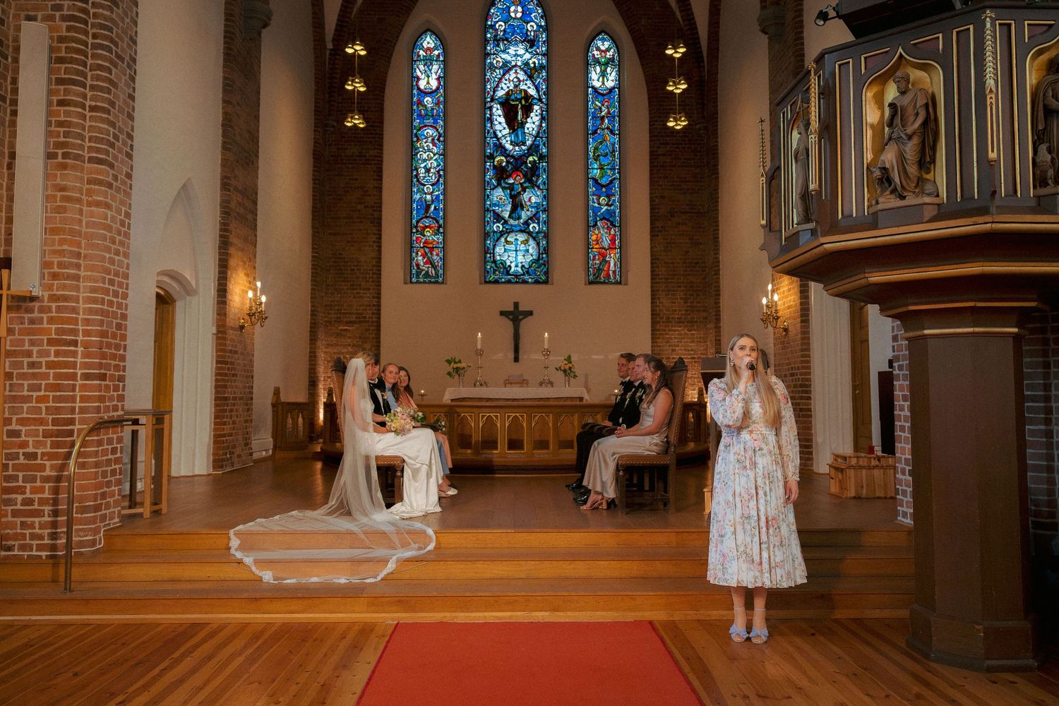 Wedding ceremony in a brick church with stained glass windows, bride seated as a singer performs.