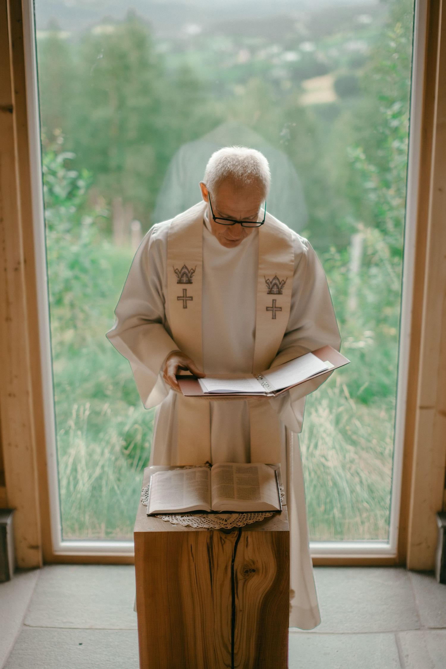 Elderly priest in white vestments reading from Bible at wooden altar before large chapel window with green landscape.