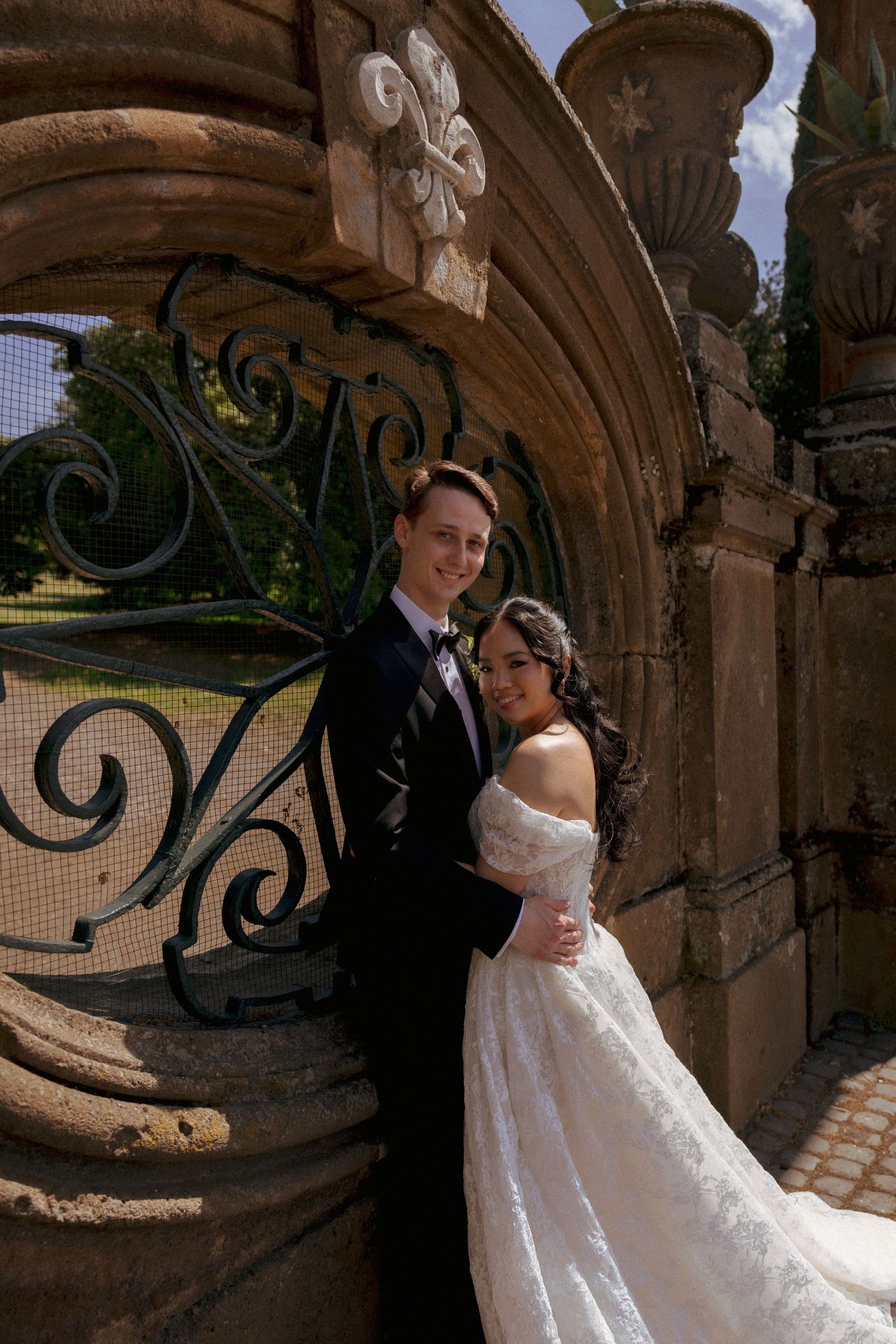 Bride and groom posing elegantly under ornate stone arch with wrought iron gate on sunny wedding day.