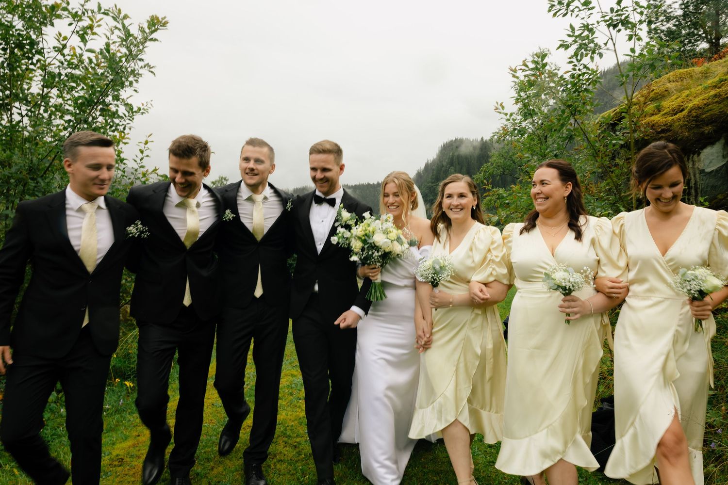 Wedding party in black suits and cream dresses walks together laughing on green lawn in Norway.