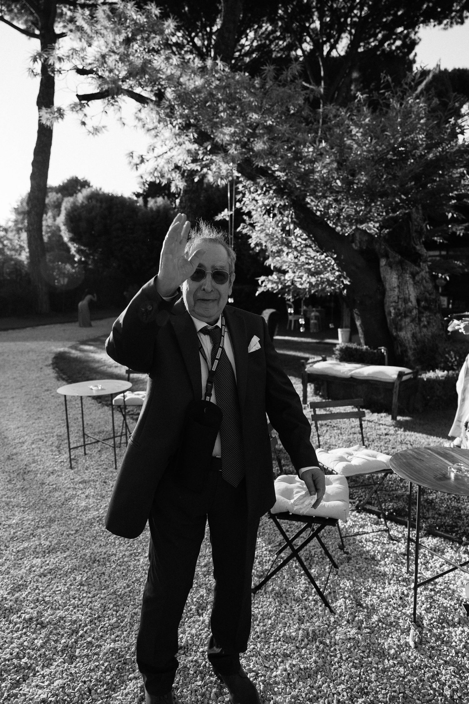 Elegant man in suit waving outdoors at a garden event, black and white photo.