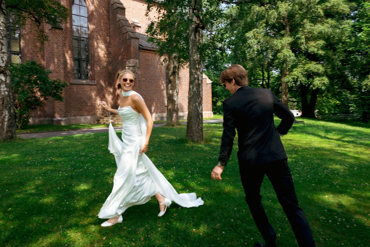 Bride in white gown and sunglasses runs playfully with groom in black suit on green lawn beside brick building.
