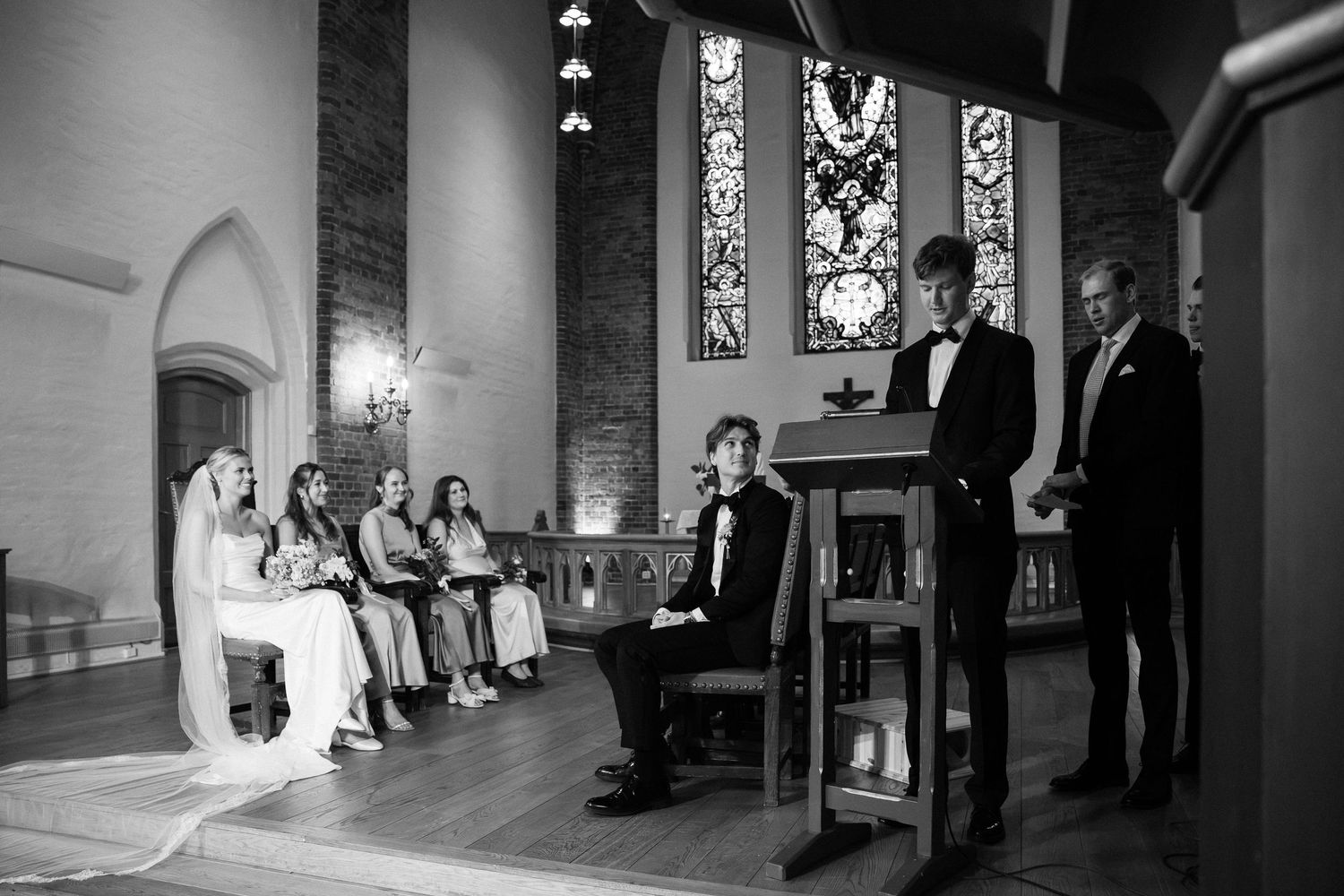 Black and white wedding ceremony photo inside a church with bride, groom, and guests seated near stained glass windows.