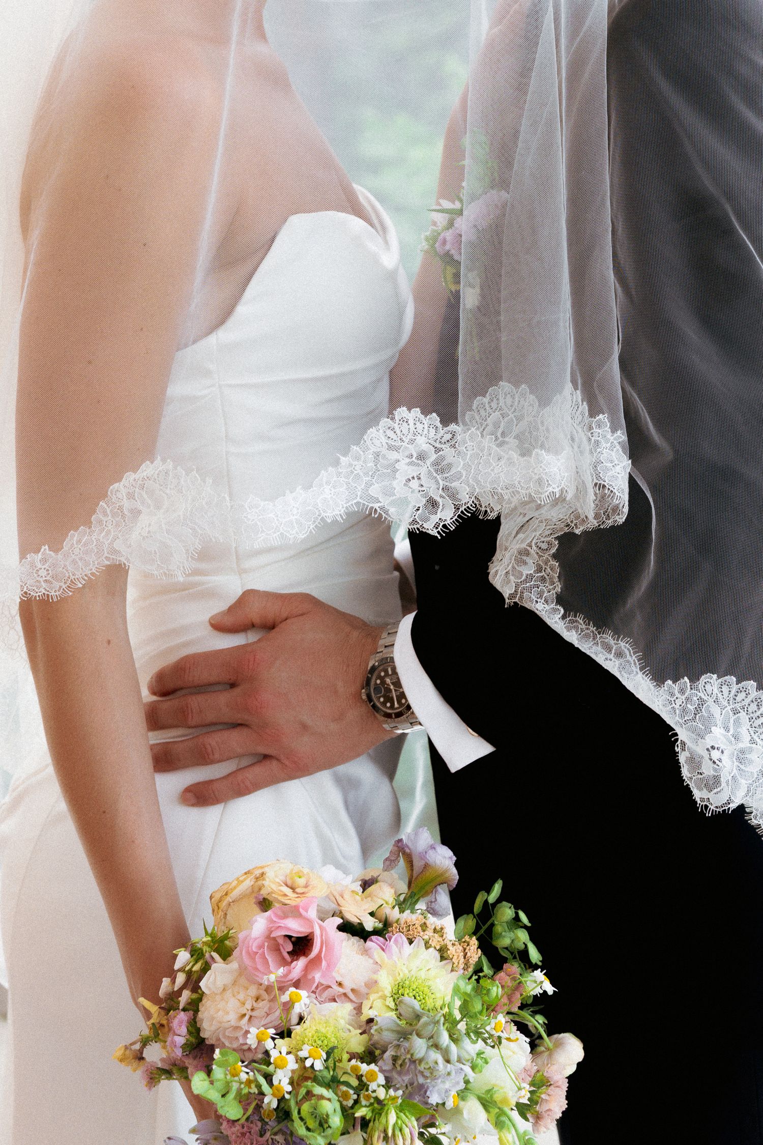 Close-up of groom placing hand on brides waist while she holds colorful wedding bouquet.
