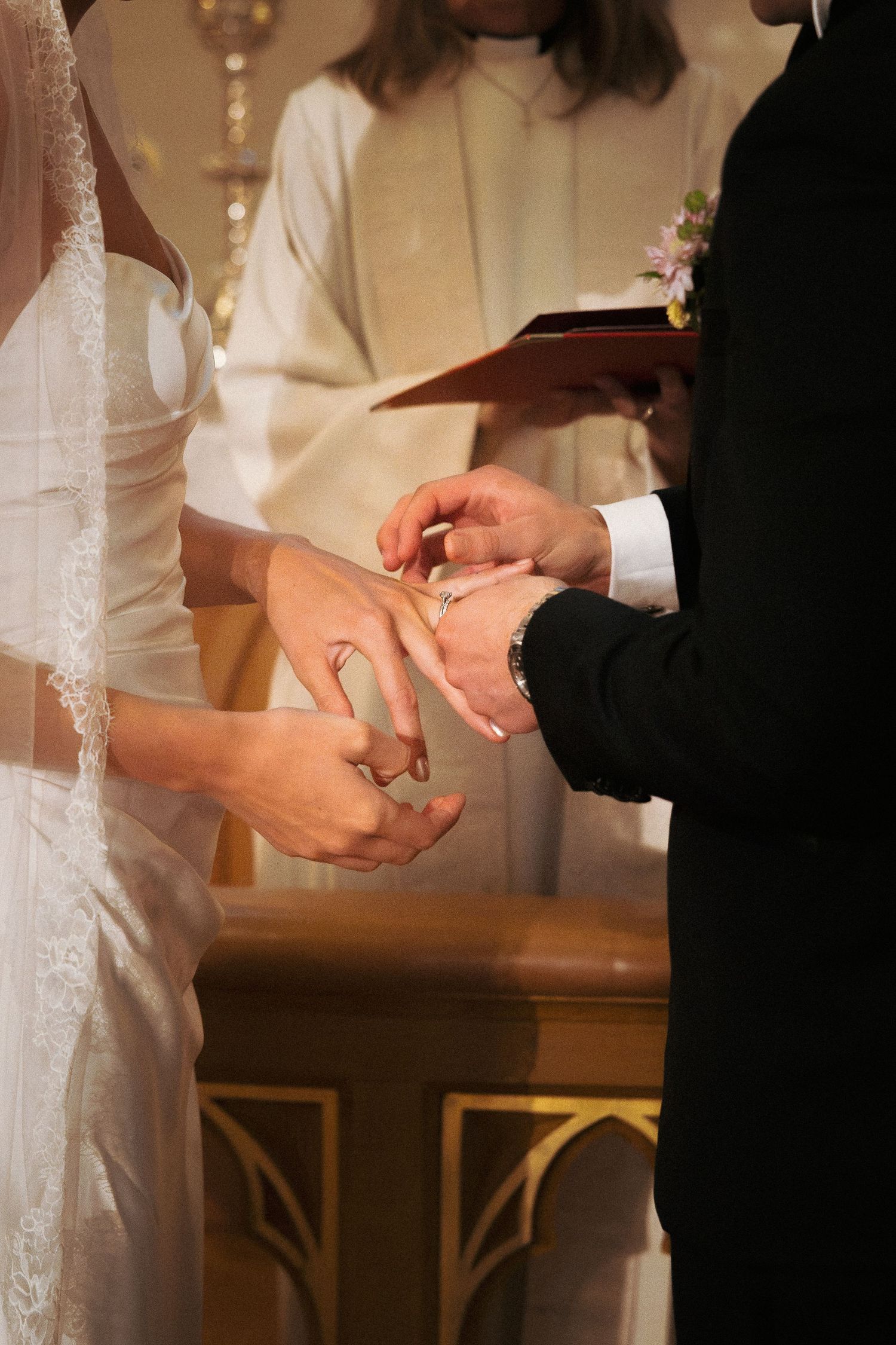 Groom placing wedding ring on bride's finger during church ceremony with officiant.