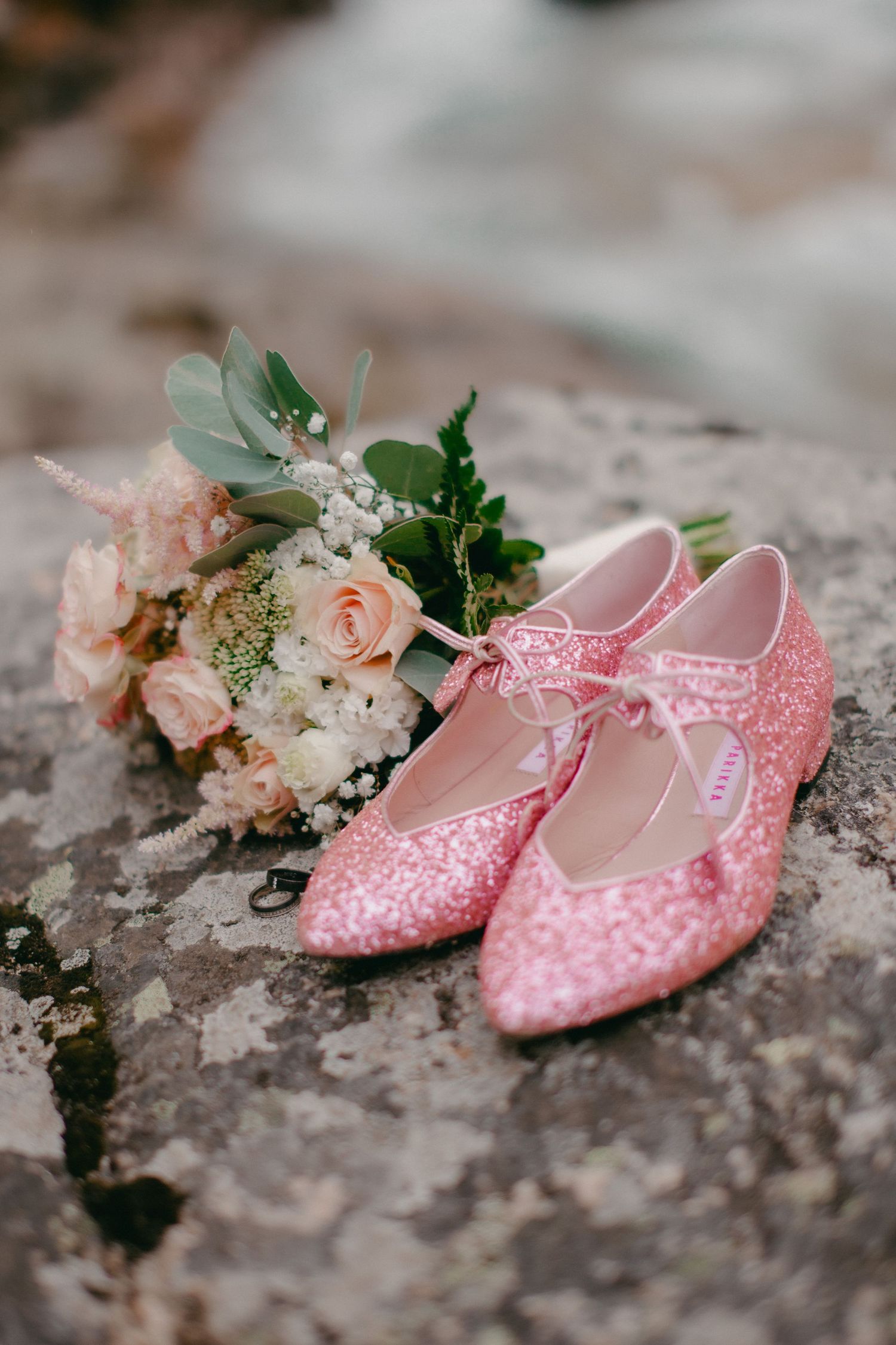 Pink glitter bridal flats with bow detail rest on a rock beside a soft pink and white wedding bouquet.