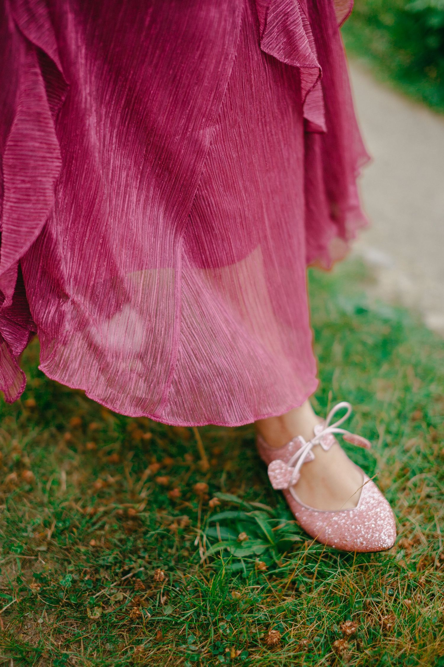 Close-up of a woman wearing pink glitter ballet flats with bow detail and a flowing mauve satin dress standing on grass.