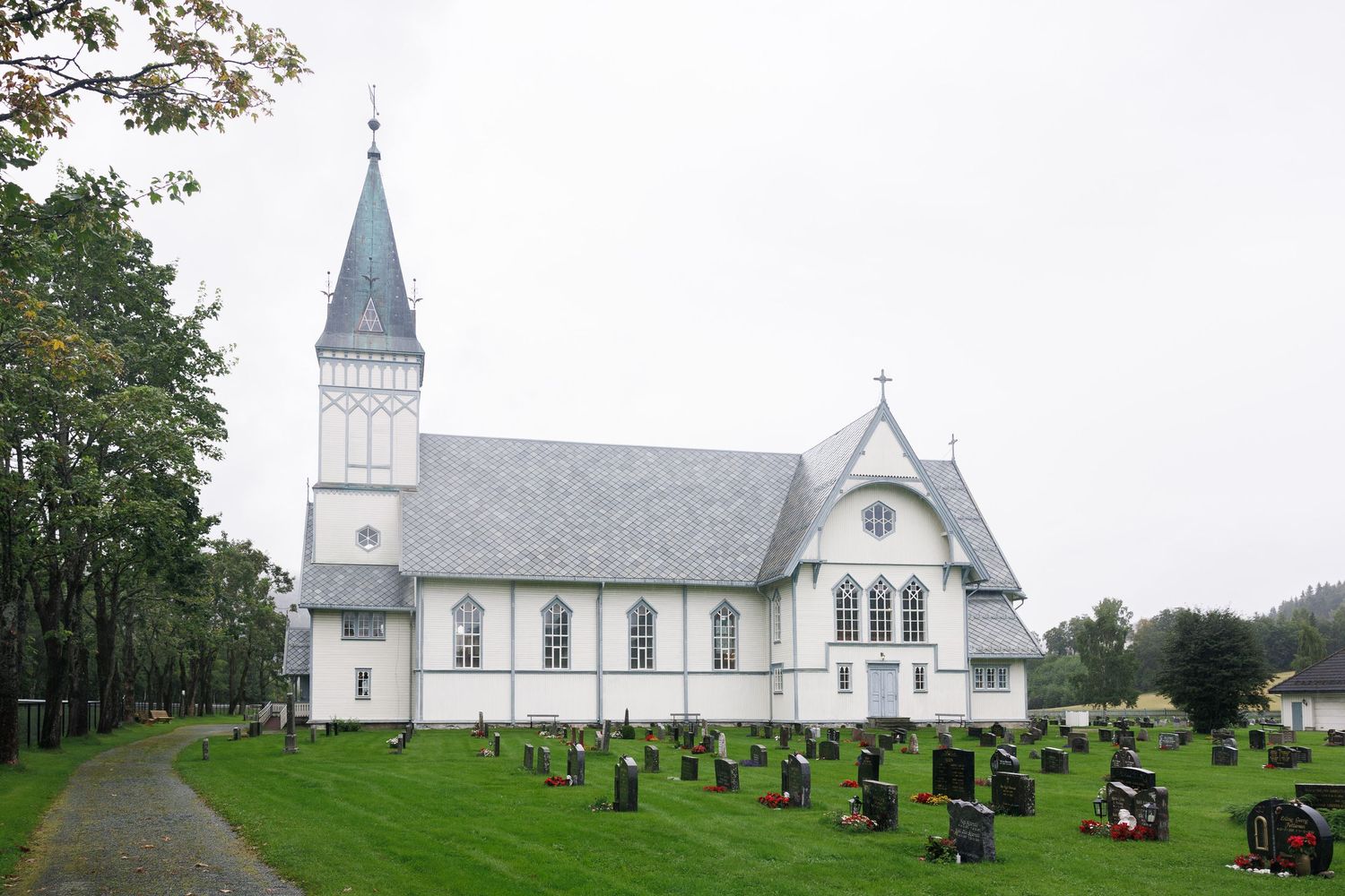 White Norwegian church with blue spire surrounded by cemetery and green trees on overcast day.
