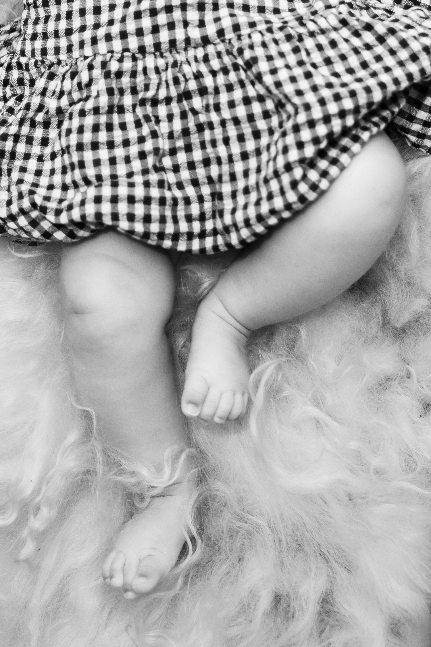 Black and white photo of a baby's chubby feet and legs in a checkered dress, resting on a fluffy fur rug.