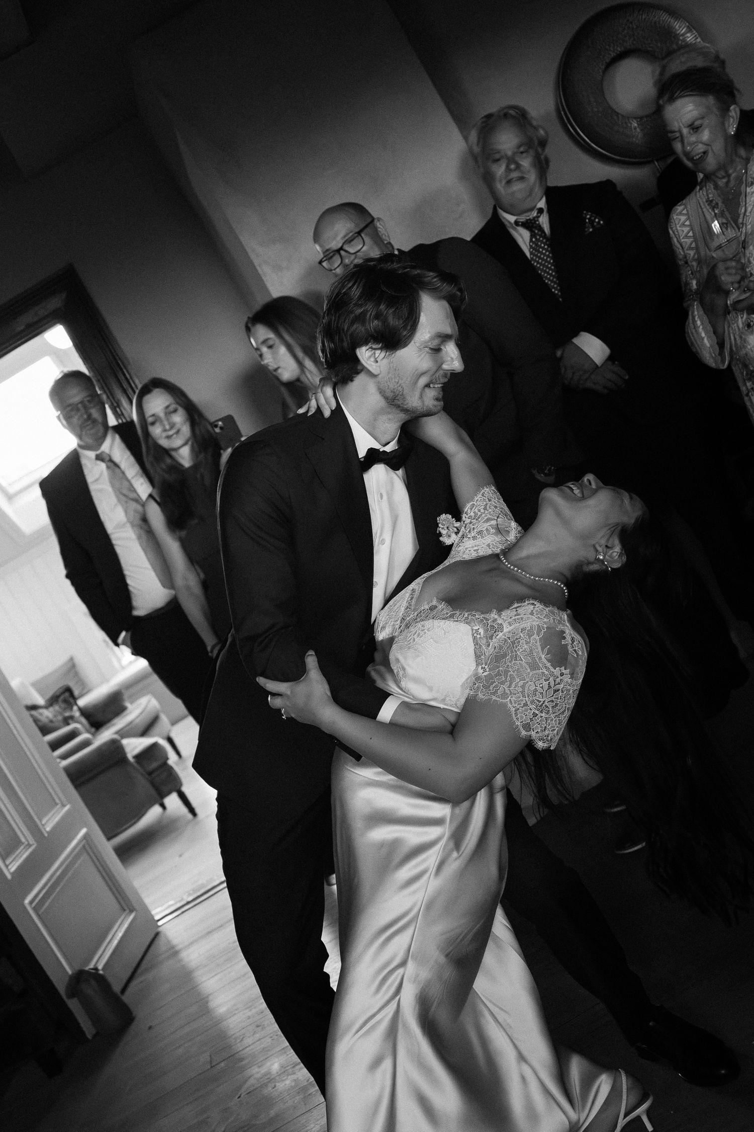 Black and white photo of bride and groom sharing first dance at wedding reception with guests watching in background.