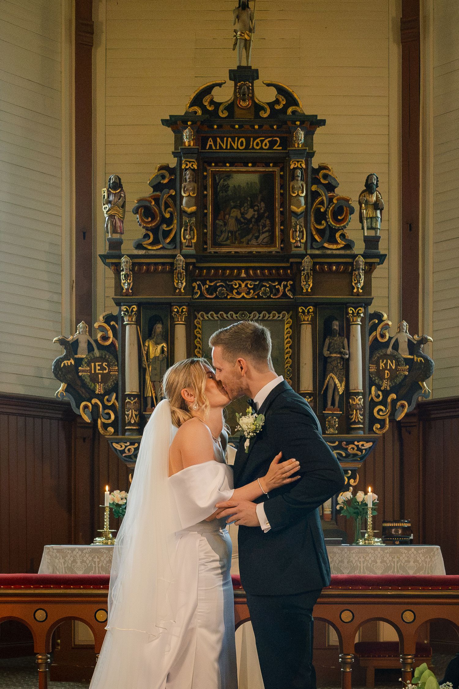 Bride and groom share a kiss at the altar of a historic church with an ornate 1662 altarpiece backdrop.