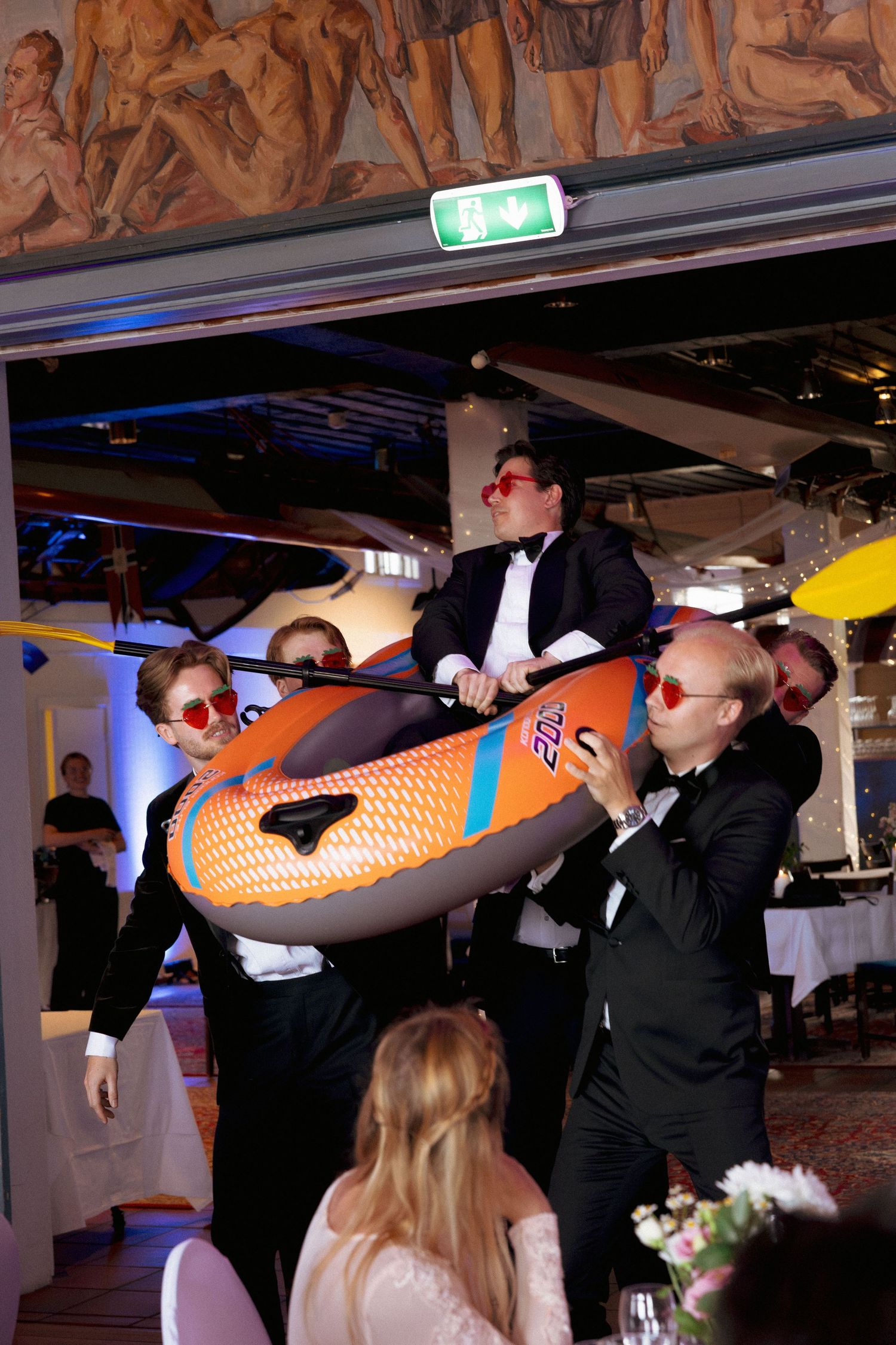 Groomsmen in tuxedos carrying an inflatable orange kayak into a wedding reception venue.