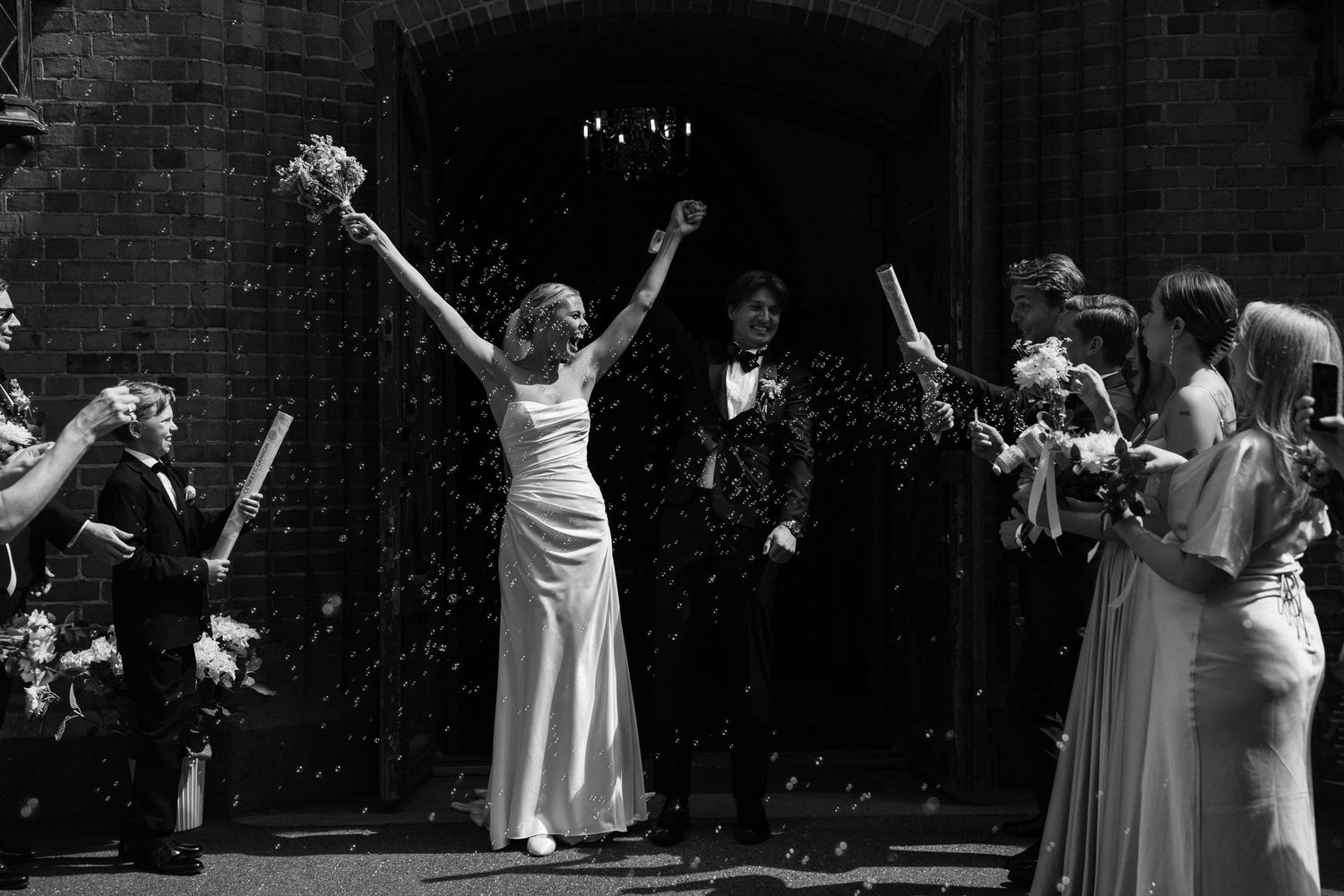 Black and white photo of a joyful bride and groom celebrating with confetti outside a brick building.