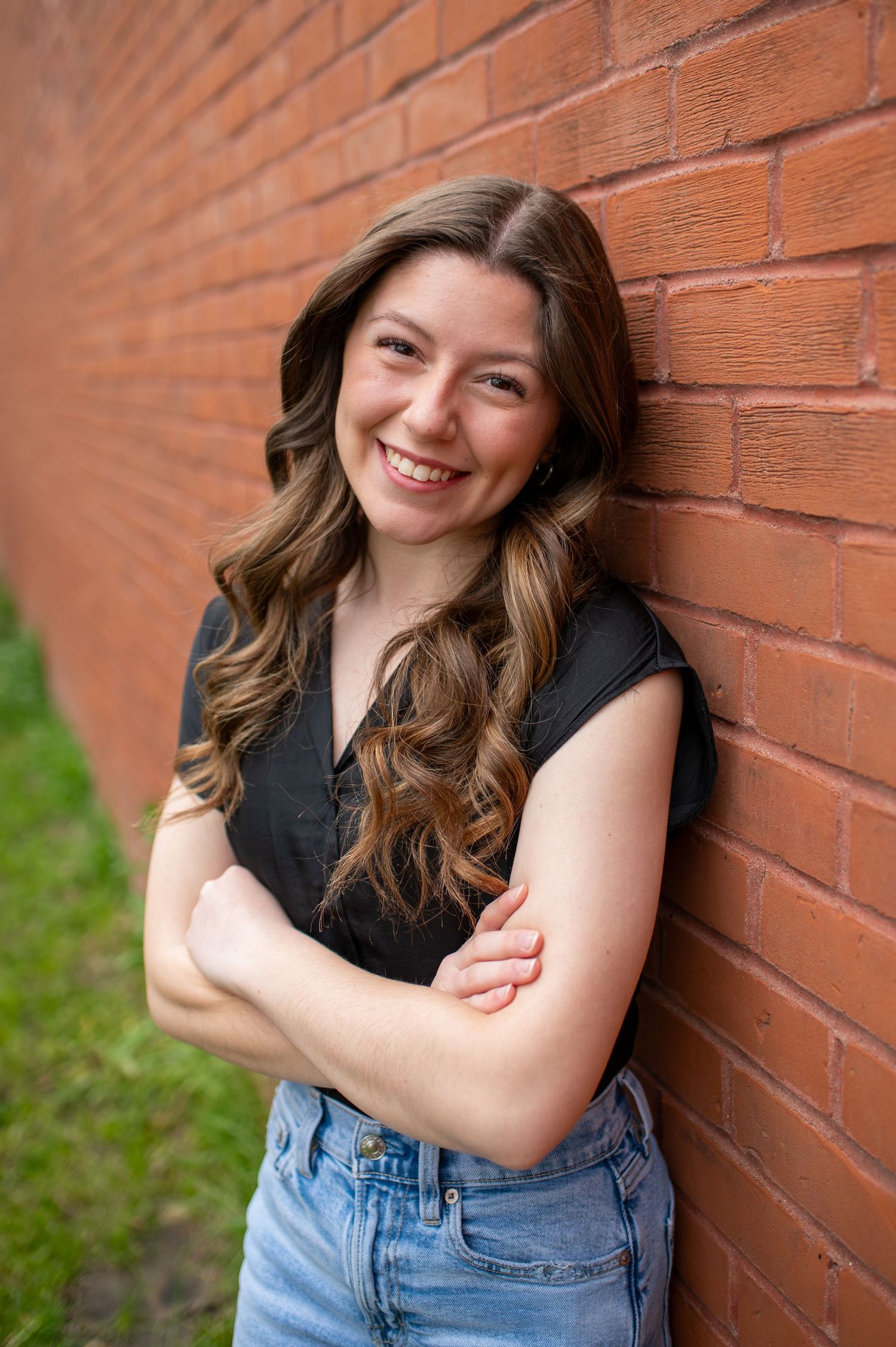 A woman in a black sleeveless top and denim jeans poses against a red brick wall outdoors for outdoor professional headshots.