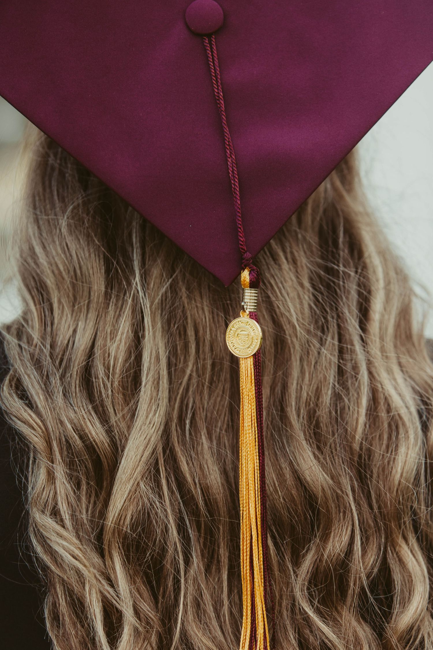 Close-up detail of burgundy graduation cap with gold tassel against wavy blonde hair.