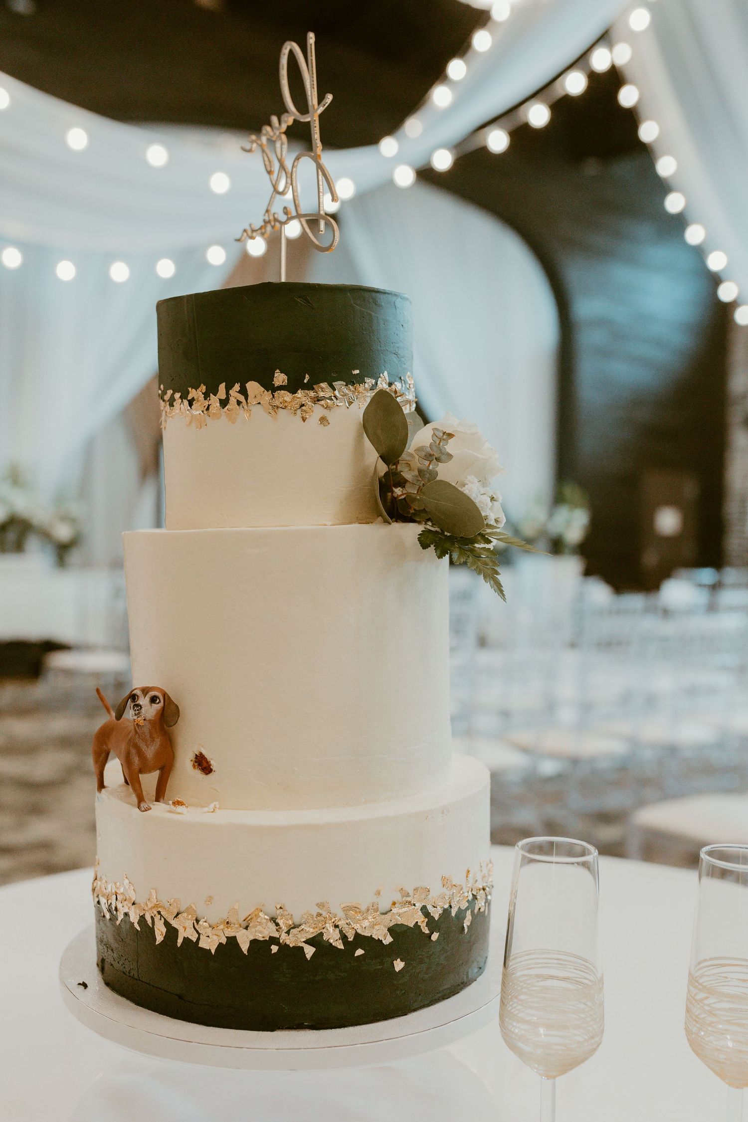 Three-tiered wedding cake with black and white design featuring metallic accents and string lights in background.