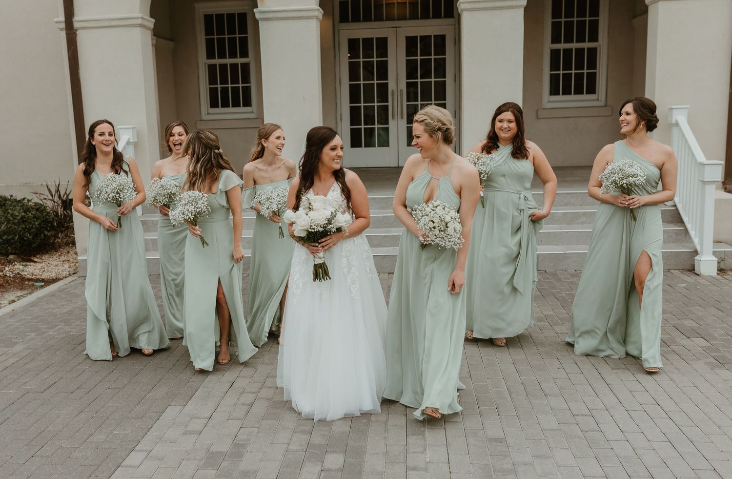 Bridesmaids in mint green dresses pose with bride in white gown outside elegant building.