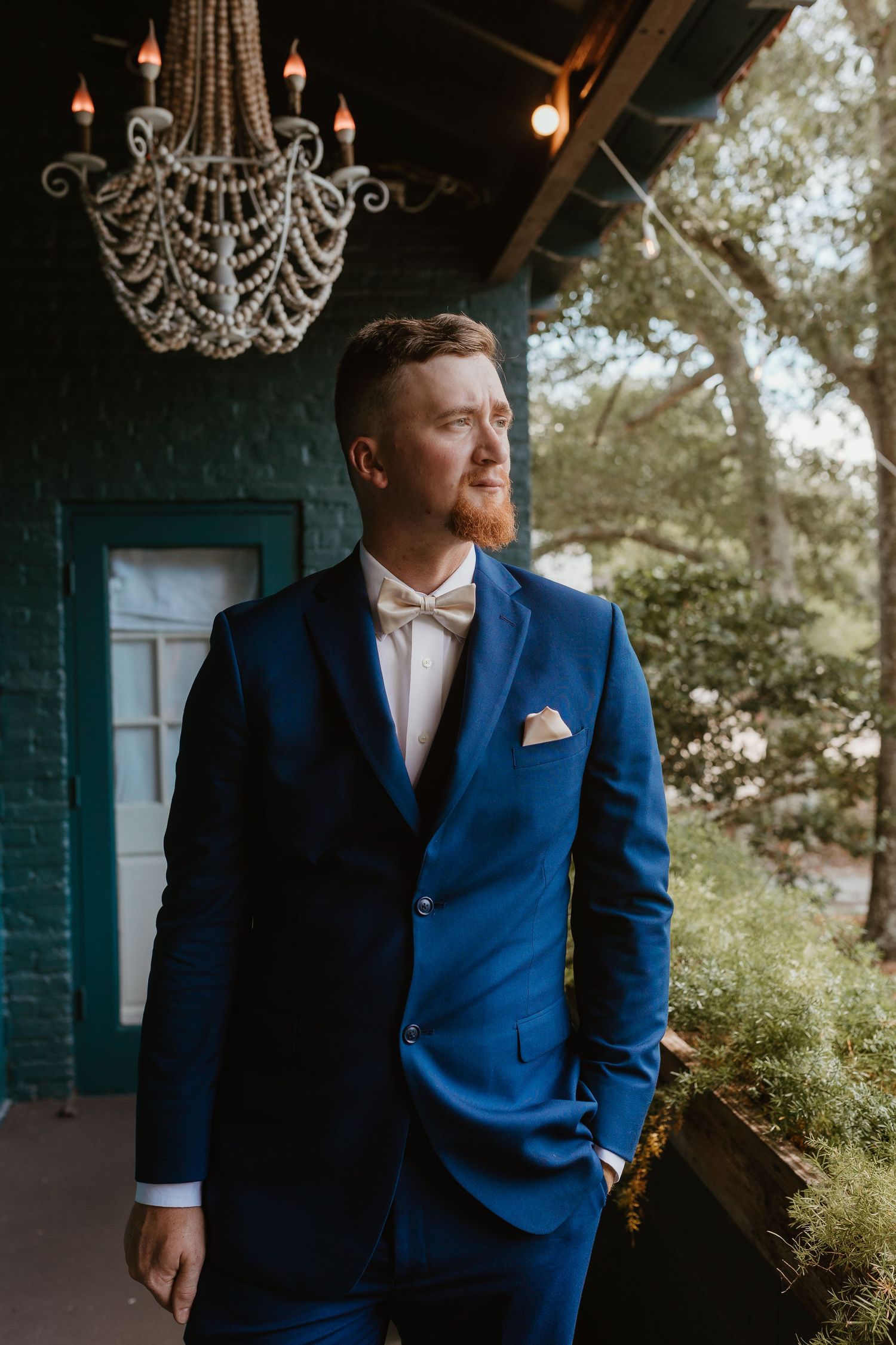 Groom in navy blue suit and bow tie standing beneath elegant chandeliers.