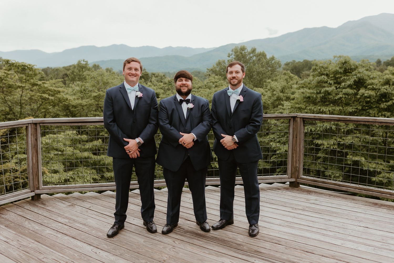 Groomsmen in dark suits posing casually on mountain view deck.