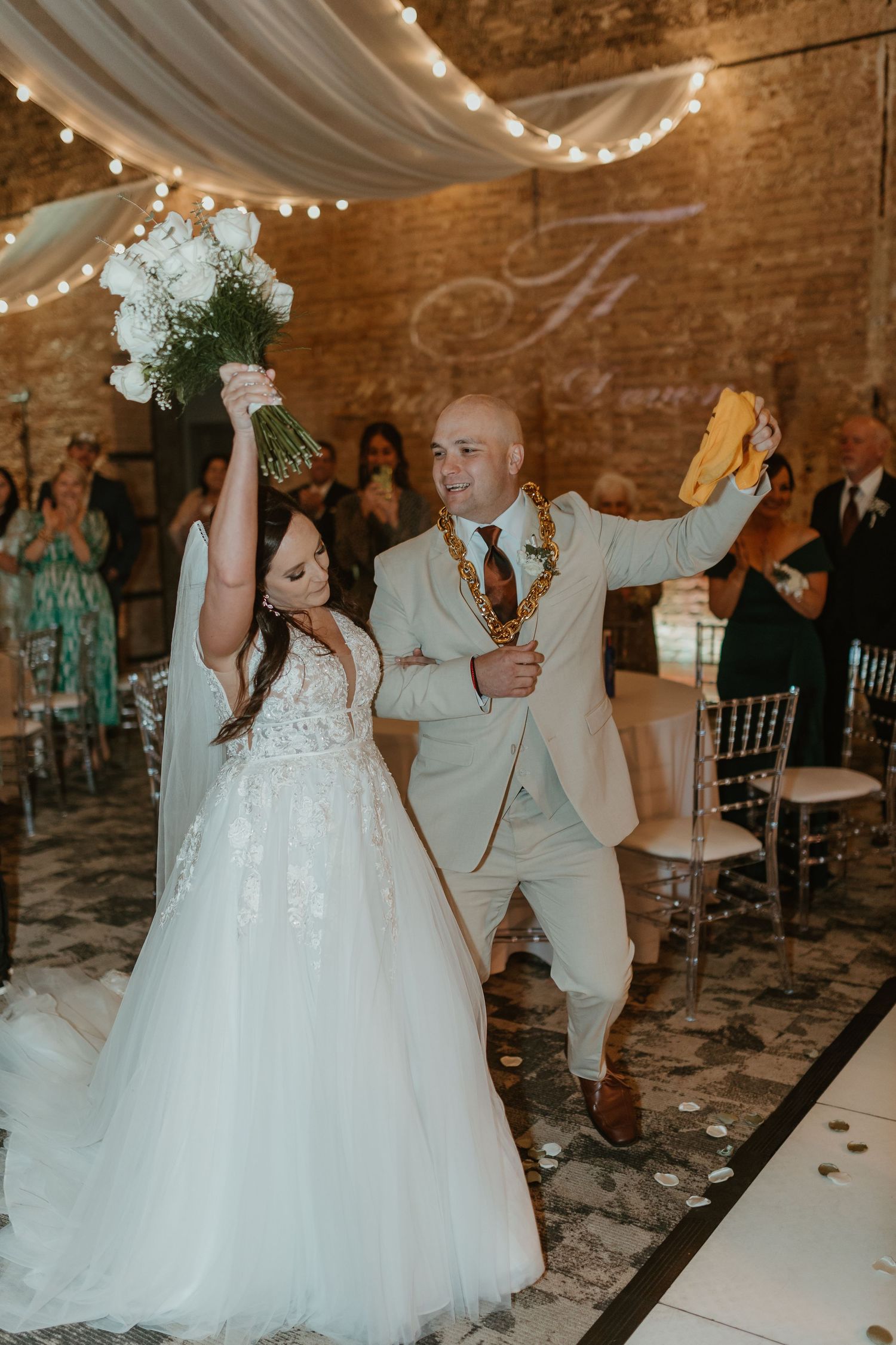 Newlyweds celebrate with raised arms during first dance in rustic venue with string lights.