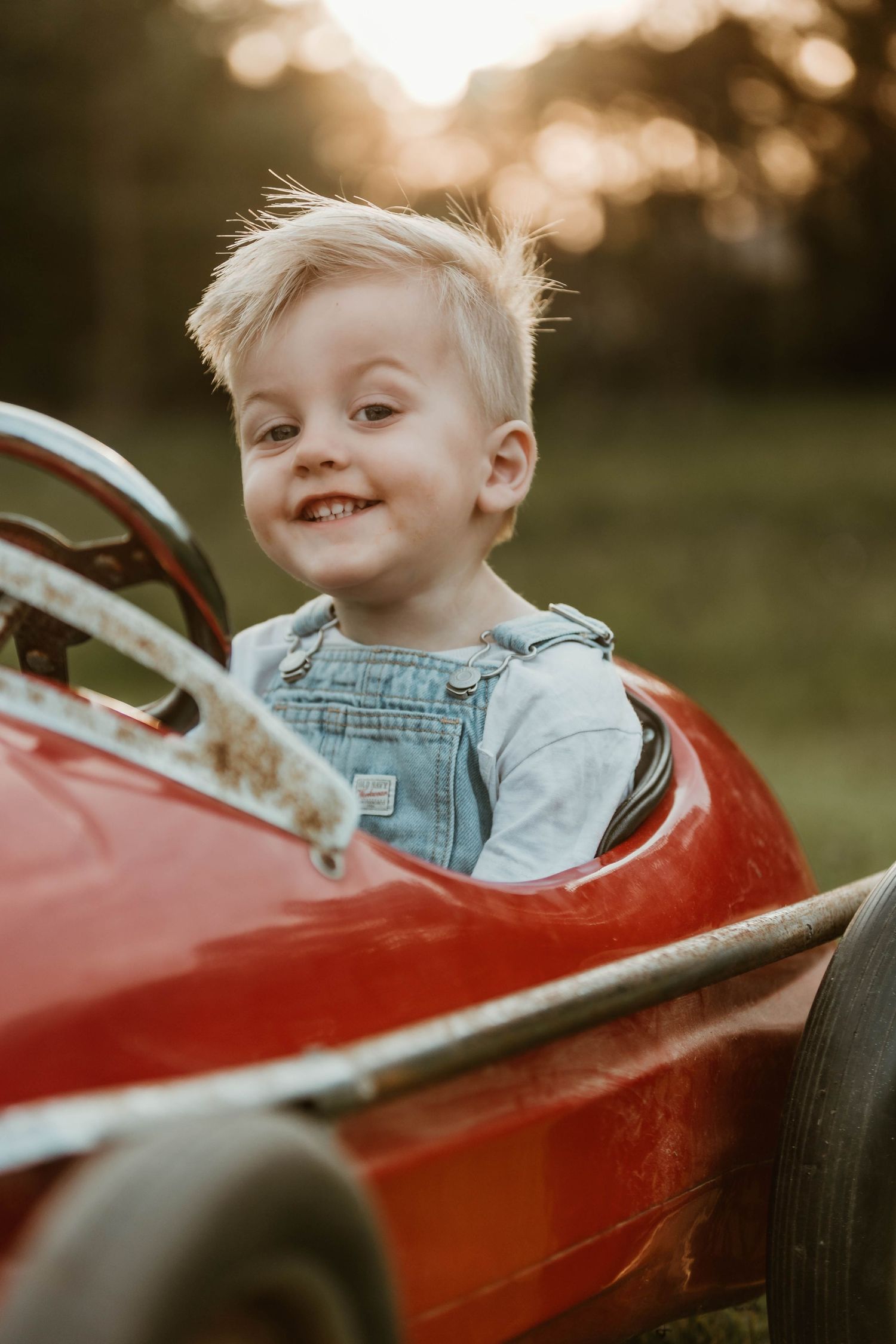 Child in denim outfit sits in classic red pedal car during sunset photoshoot.