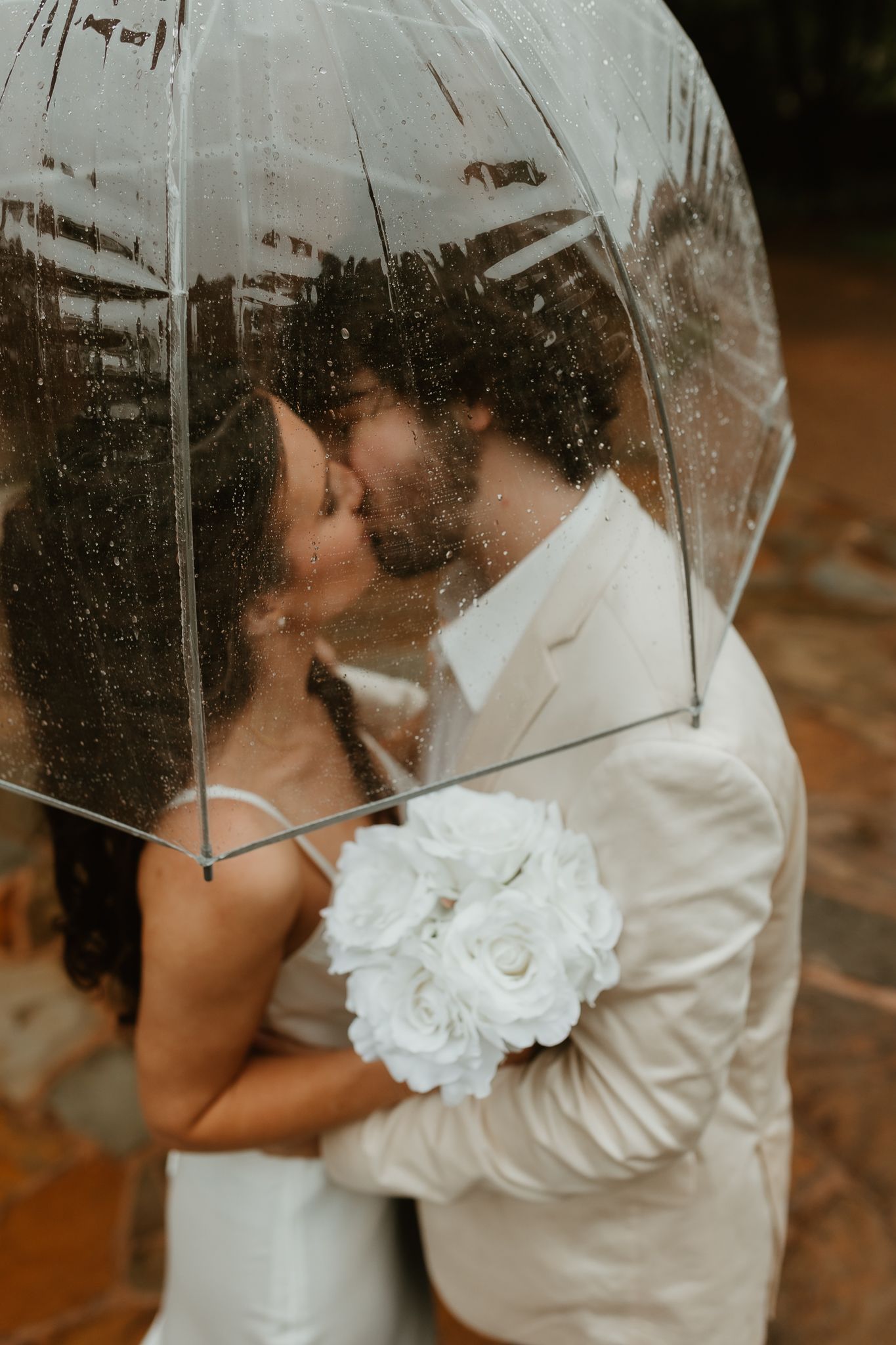 A romantic rainy day wedding photo under a clear umbrella with white floral details.