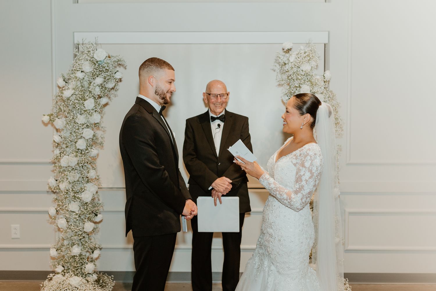 Wedding ceremony taking place in a modern white venue with floral arch decorations.