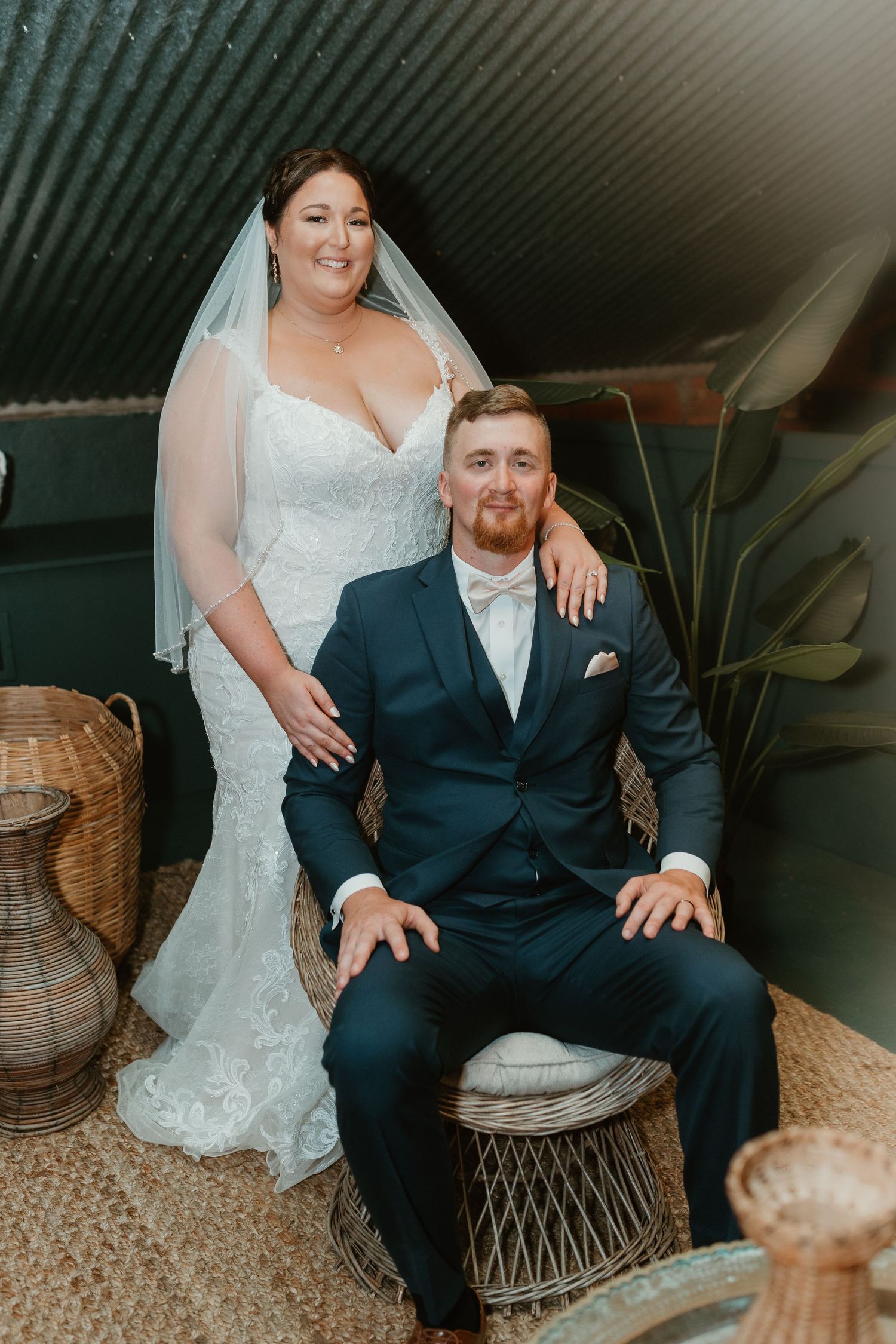Wedding portrait of couple in elegant attire seated in rustic indoor setting.