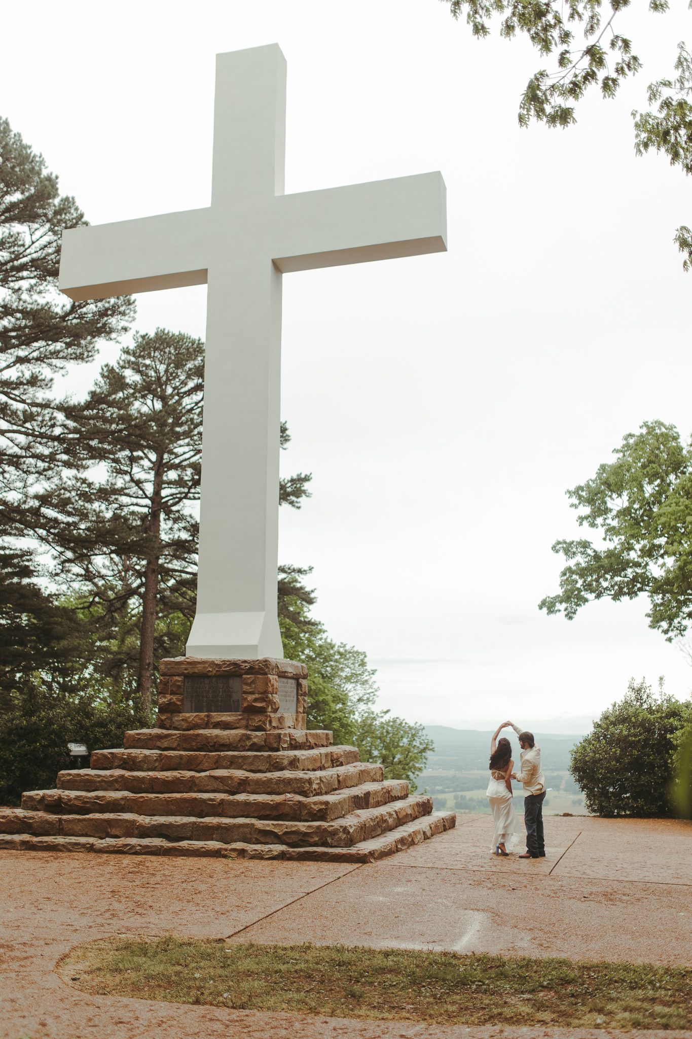 Stone steps lead up to a white cross monument overlooking a scenic mountain landscape.