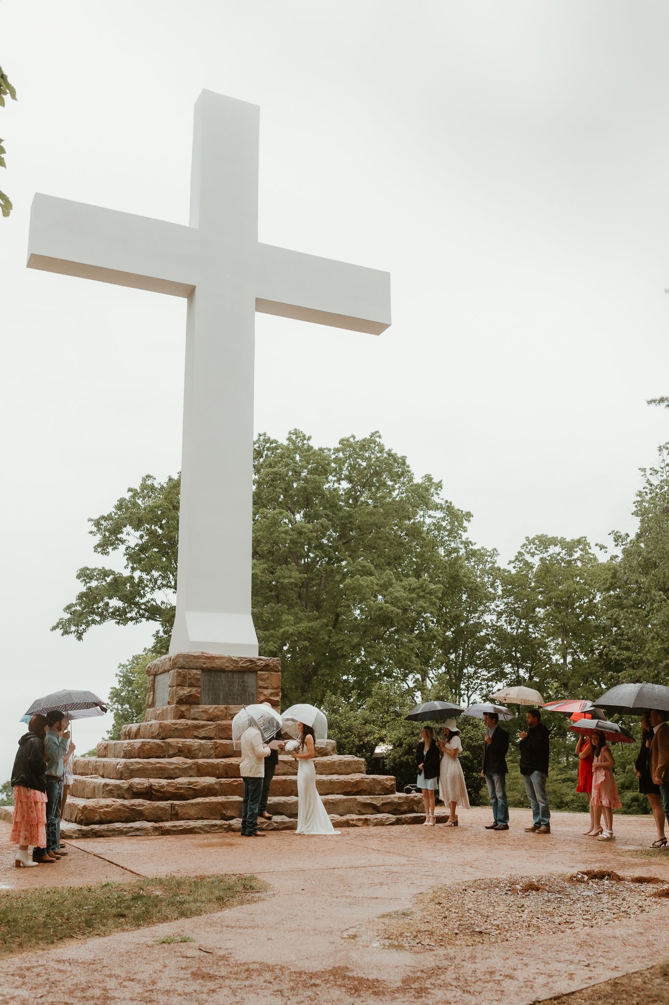 Large white cross stands atop stone platform during outdoor wedding ceremony.