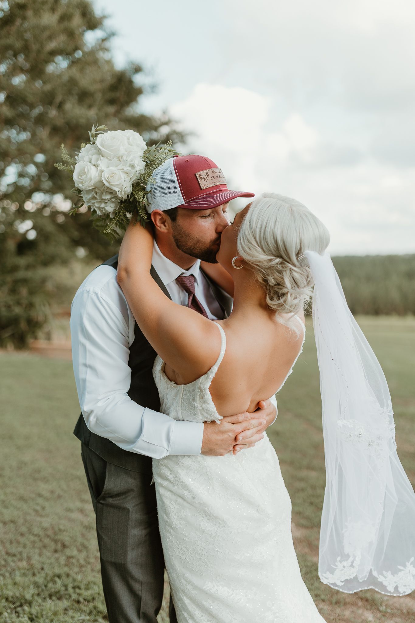 A romantic wedding couple sharing a kiss outdoors, with the bride in a white dress and groom wearing a red baseball cap.