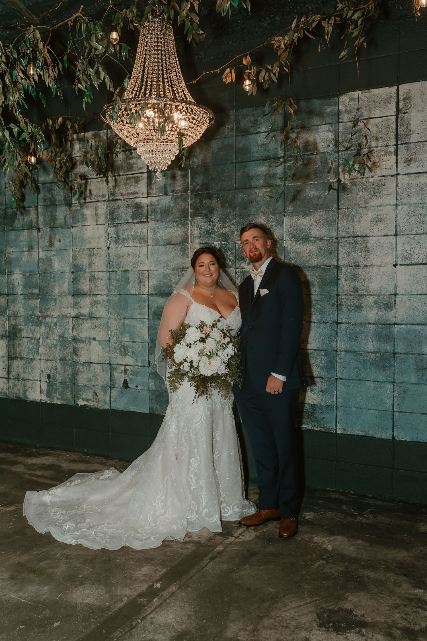 A wedding couple poses beneath an elegant crystal chandelier against an industrial blue-gray wall.