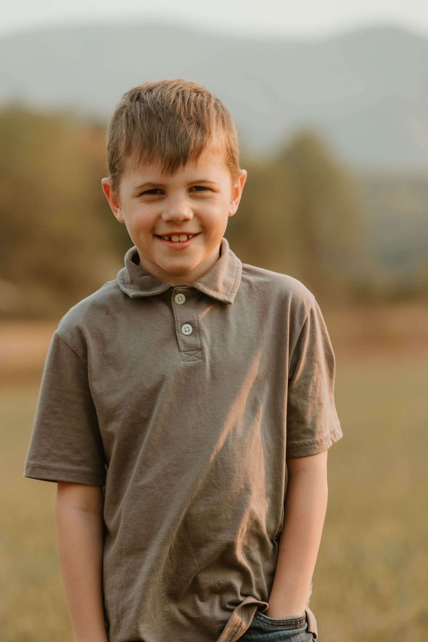 Young child wearing brown polo shirt smiling outdoors in a field during golden hour.