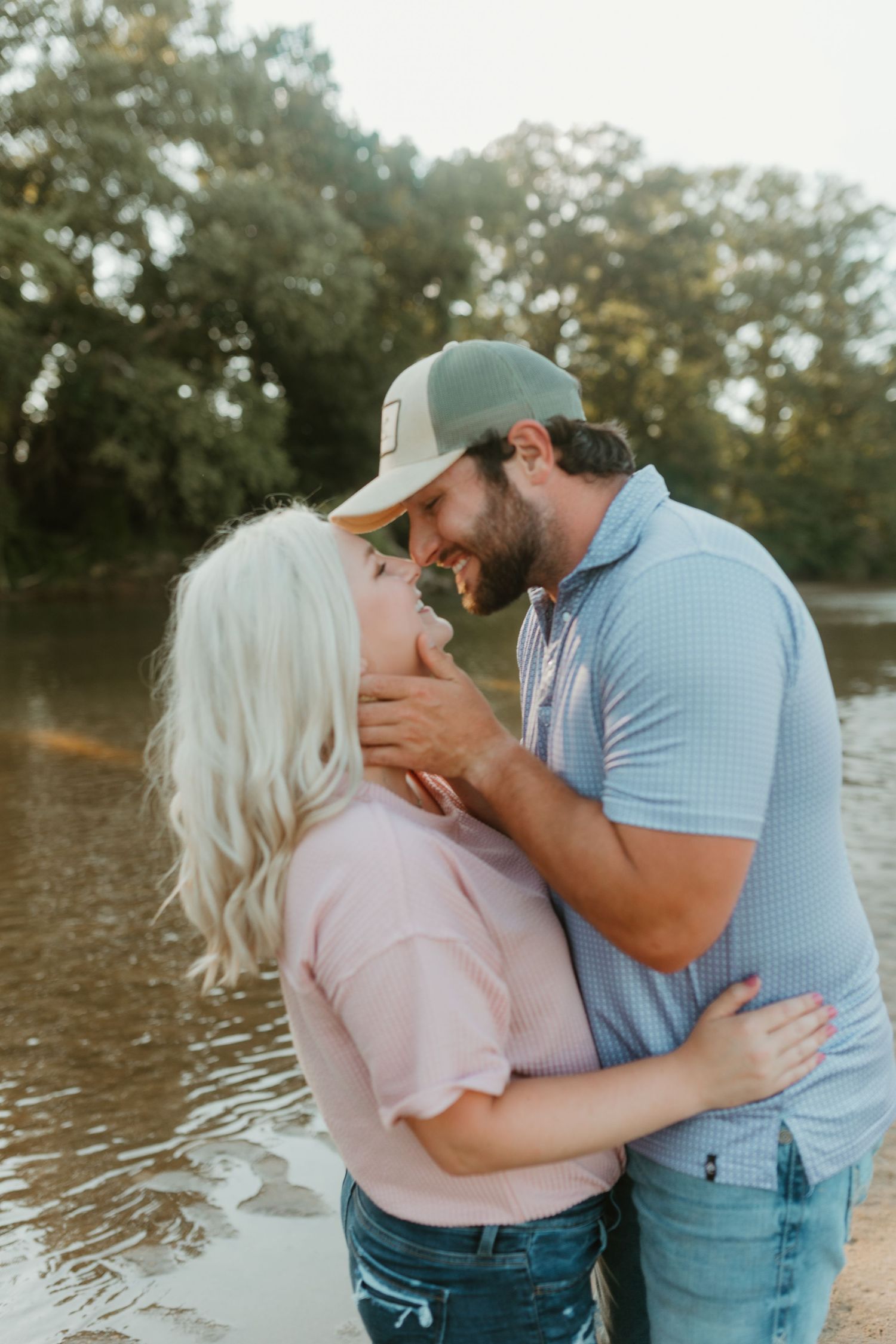 Romantic couple embracing by a lake at sunset with warm golden light filtering through trees.