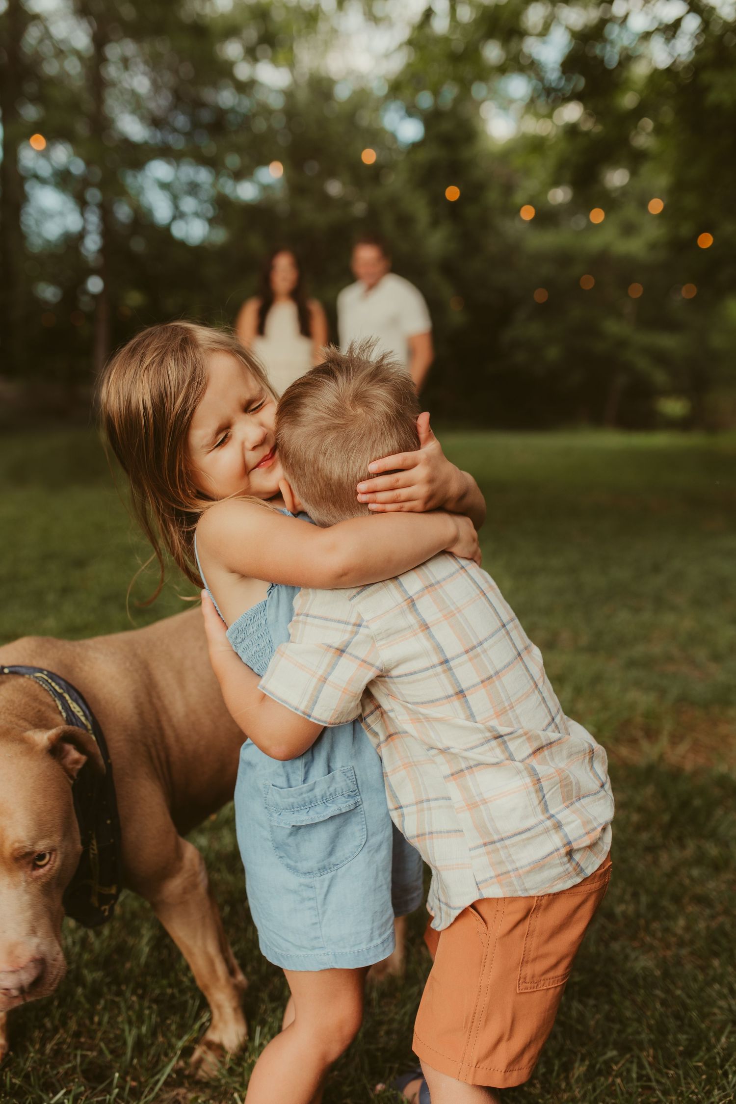 Two young children share a sweet embrace while playing outdoors at sunset with string lights in the background.