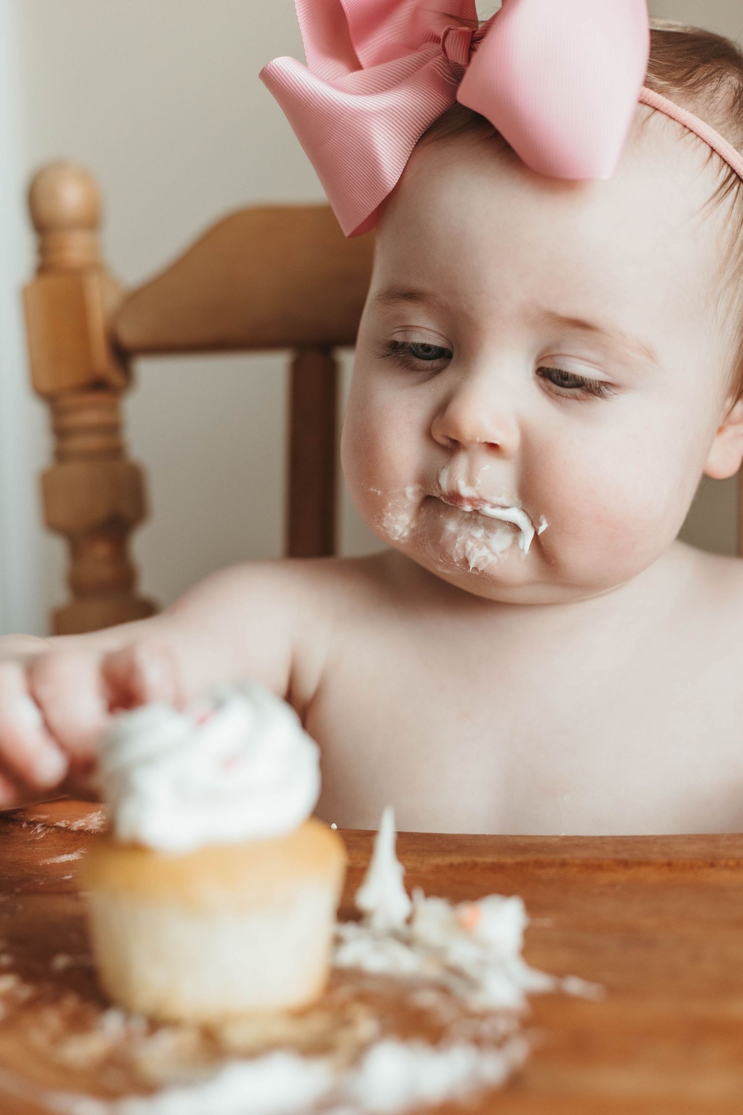 Baby with pink bow gets messy eating white frosted cupcake.