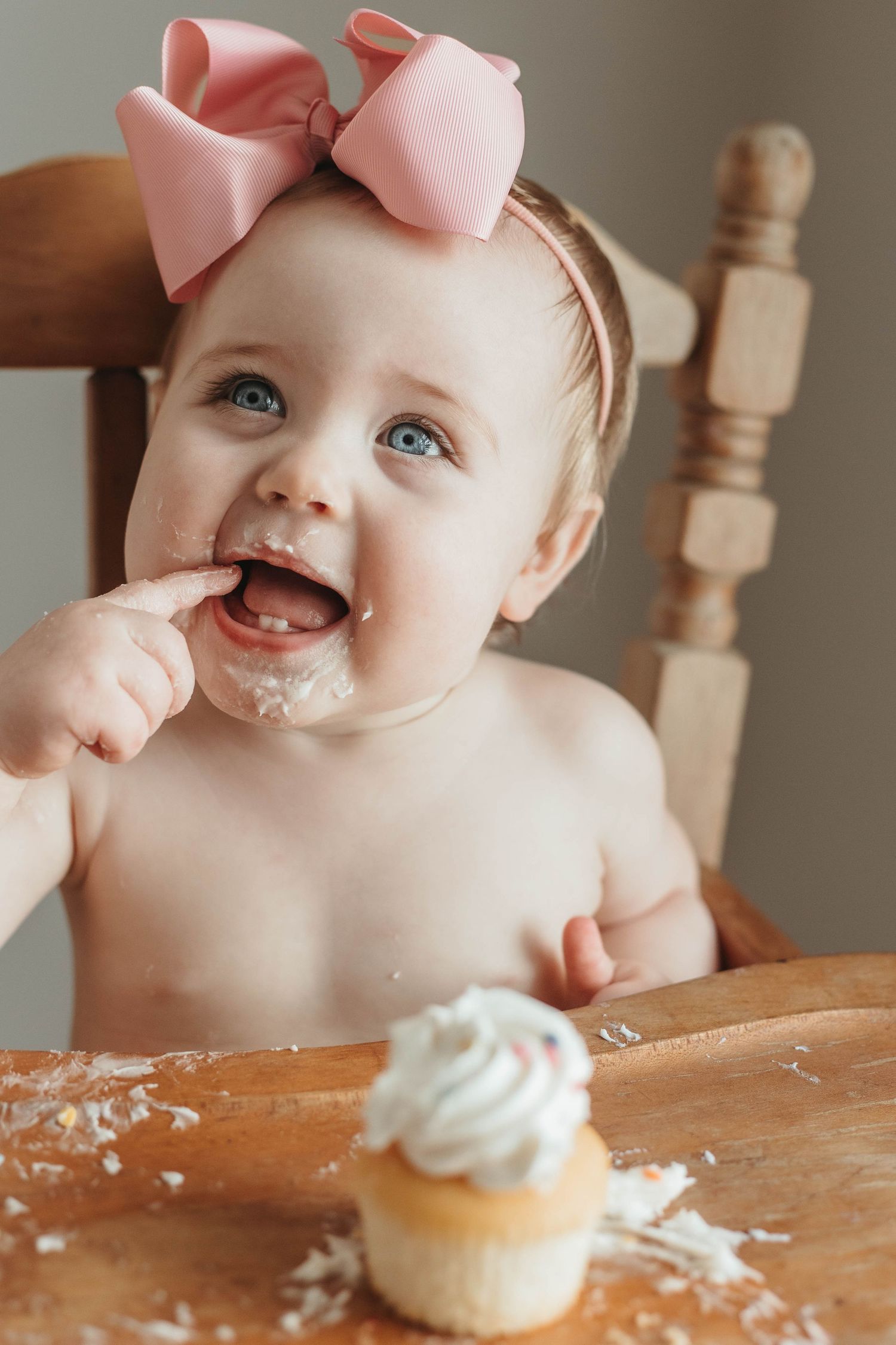 Baby wearing pink bow enjoys first birthday cupcake while sitting in wooden chair.