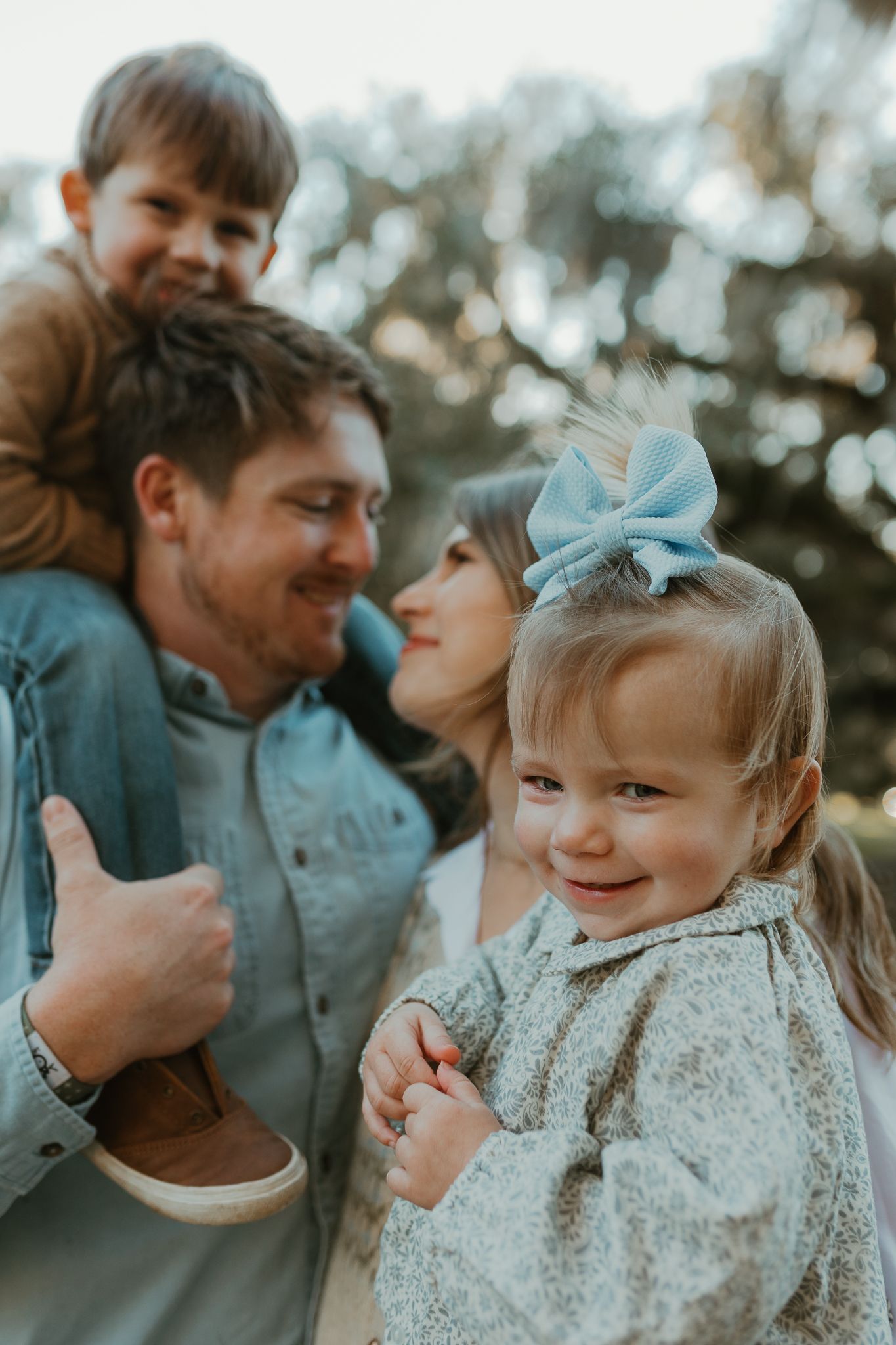 Candid family moment outdoors with playful expressions and a little one wearing a blue bow.
