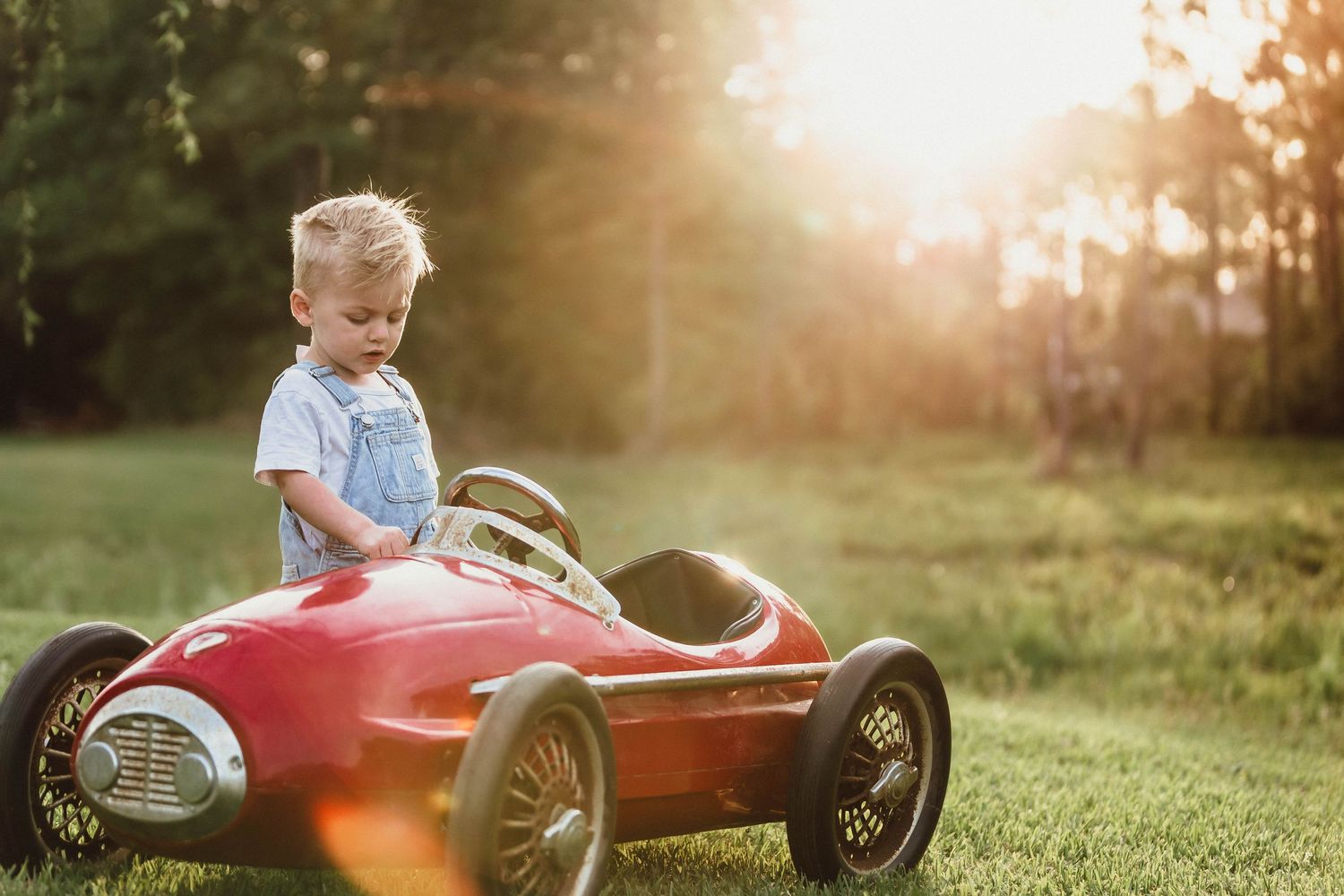 Vintage red pedal car sits in grassy field during golden hour sunset.