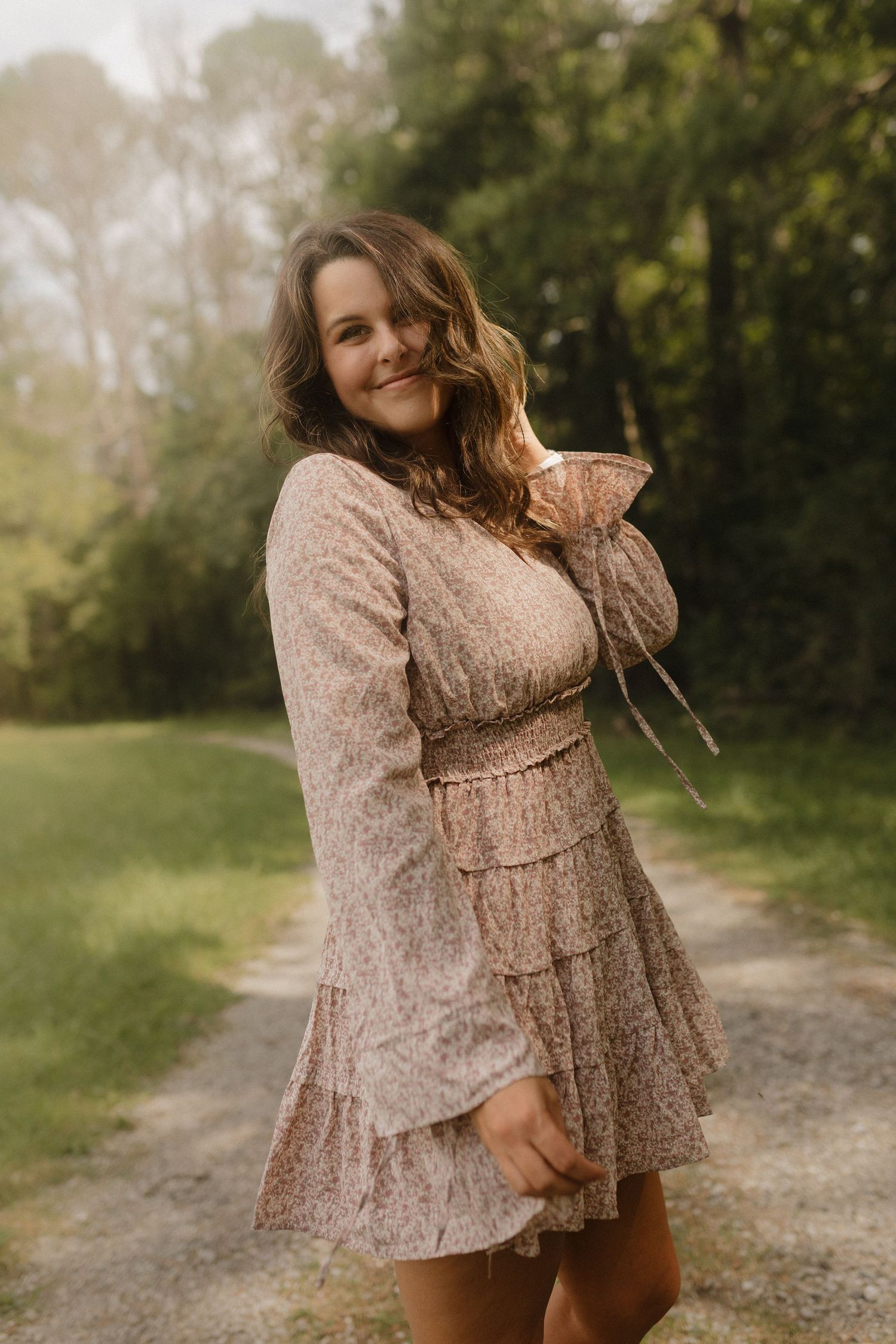 A person in a flowy beige floral dress poses playfully on a dirt path surrounded by greenery.