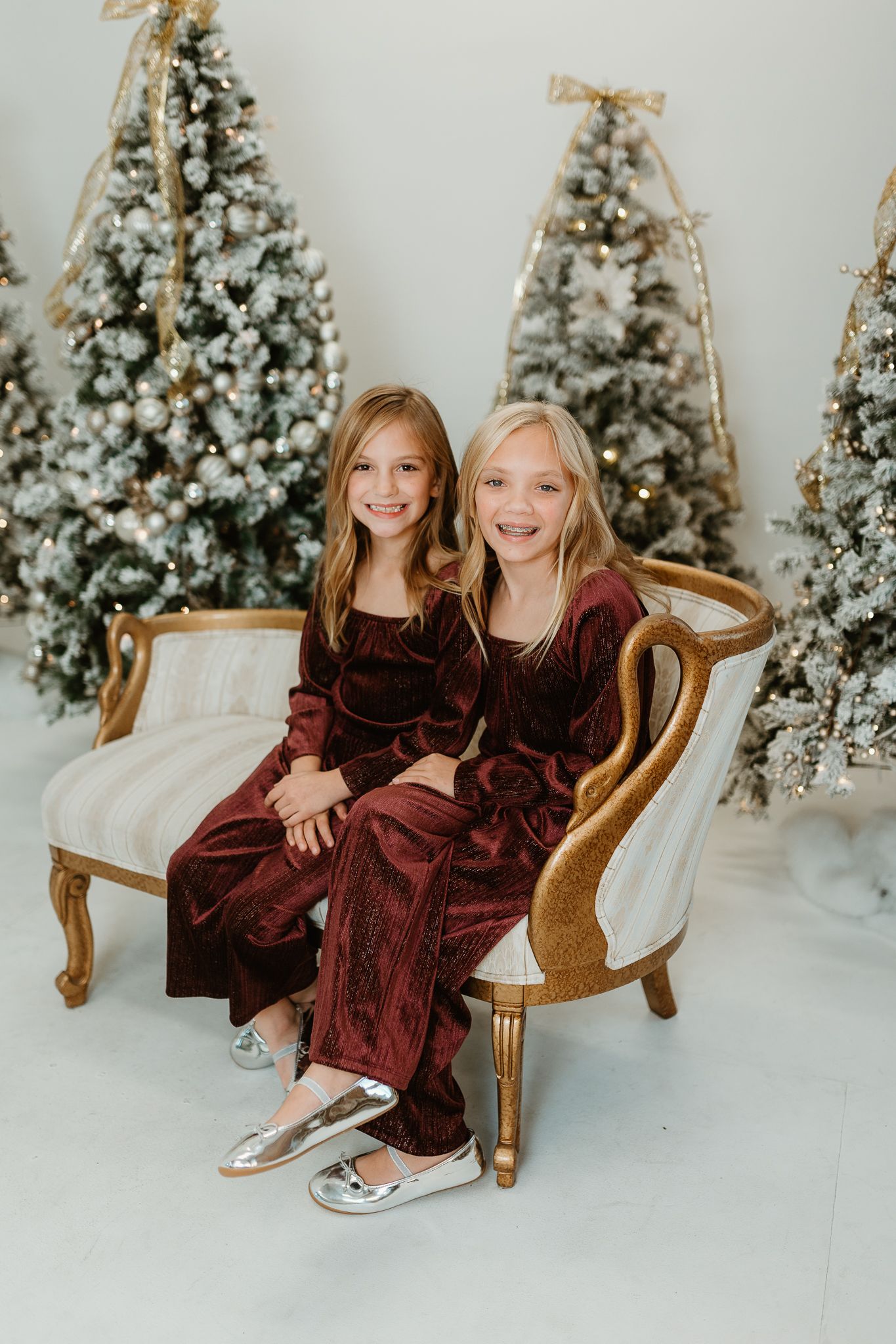 Two young girls in burgundy velvet dresses sitting on an antique sofa surrounded by snowy Christmas trees.