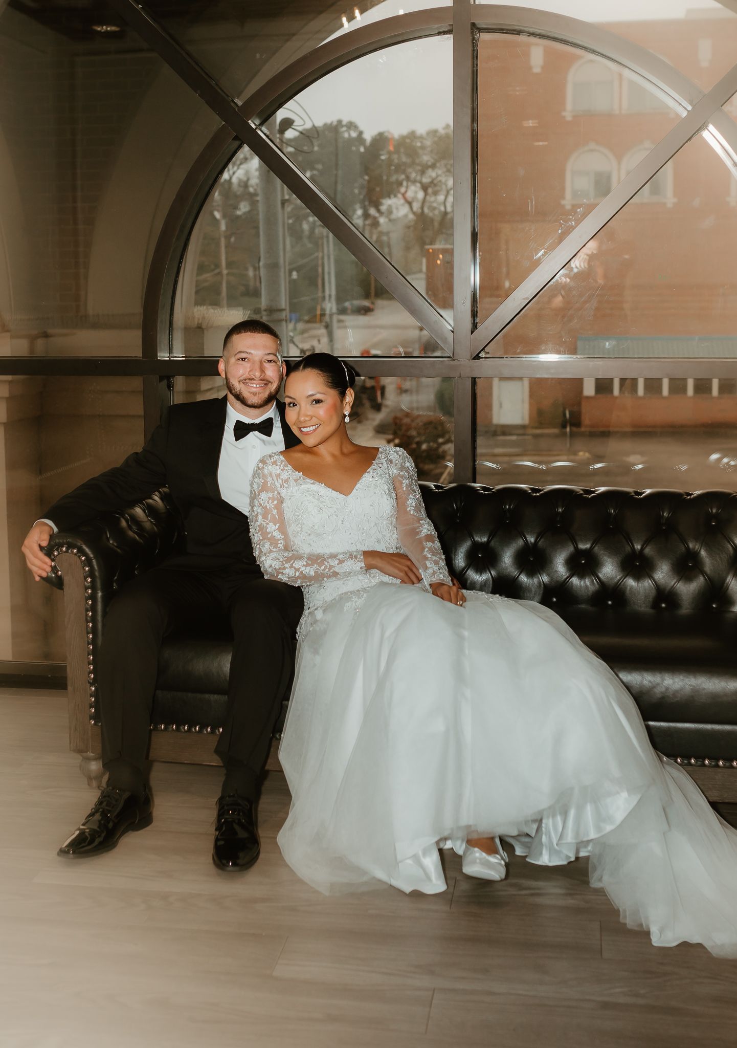 Wedding couple relaxing on leather couch beneath arched window in elegant indoor setting.