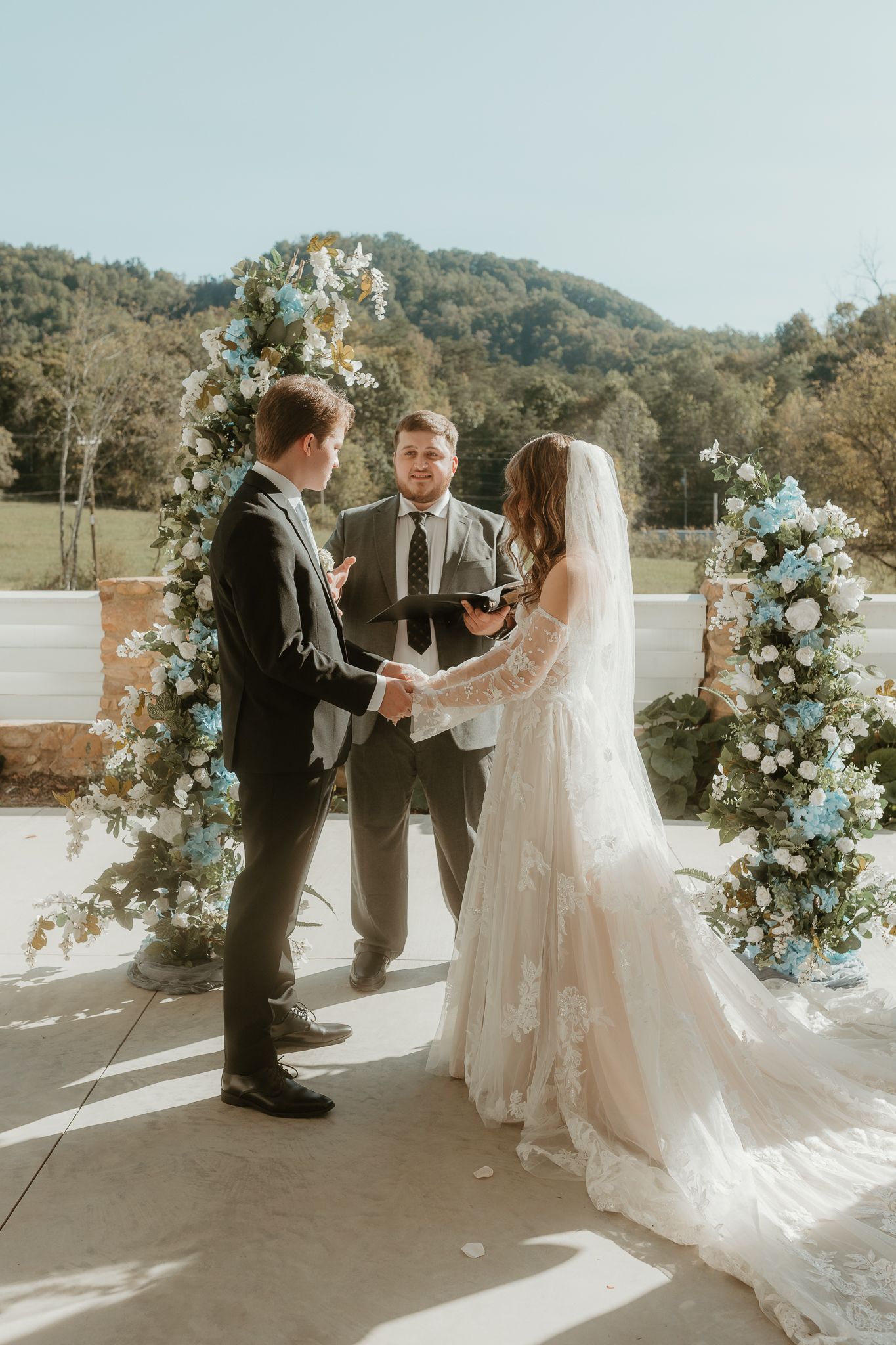 Wedding ceremony taking place under white archway decorated with blue and white flowers.